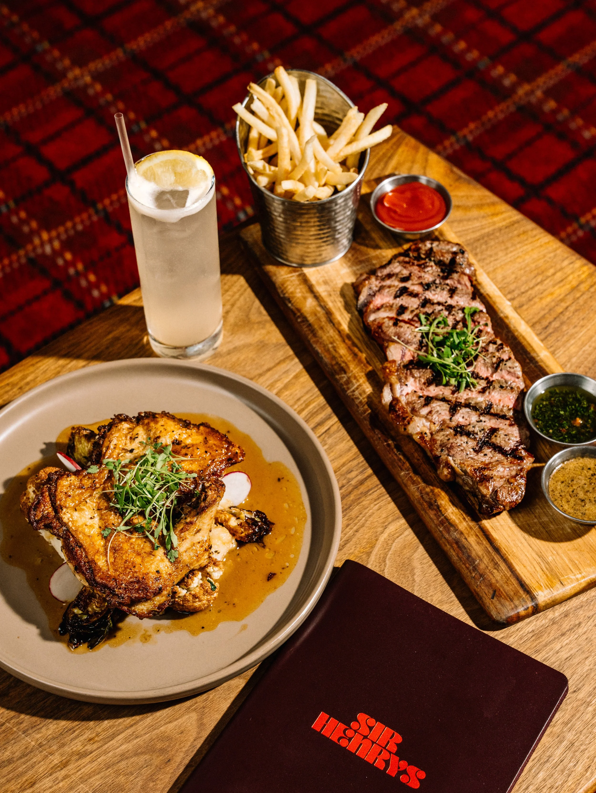 A dining table with a steak, a roasted chicken thigh in sauce, French fries, ketchup, and three dipping sauces, with a lemon drink in a tall glass.