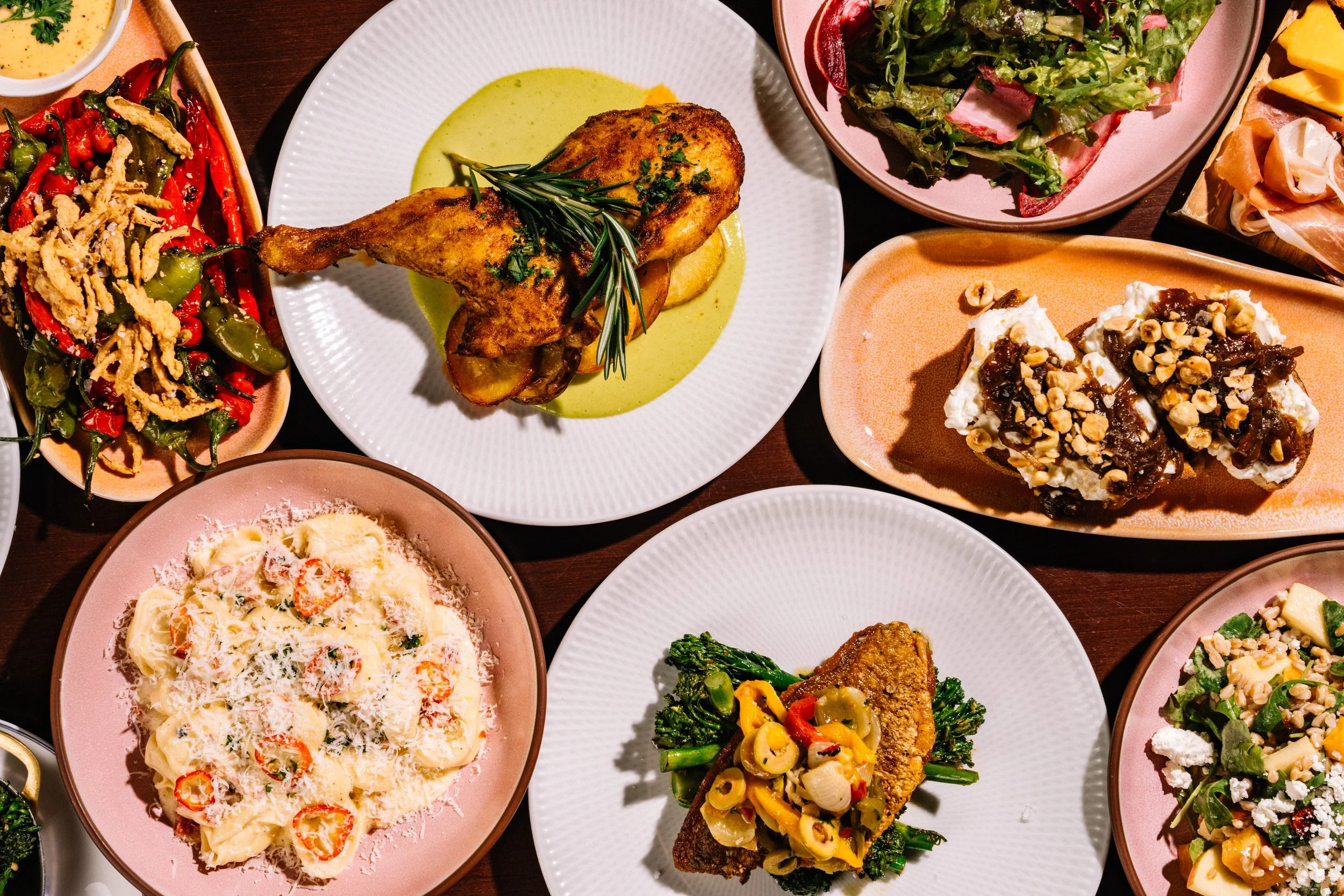 A variety of plated dishes including roasted chicken, pasta, salads, and desserts arranged on a dark wooden table.
