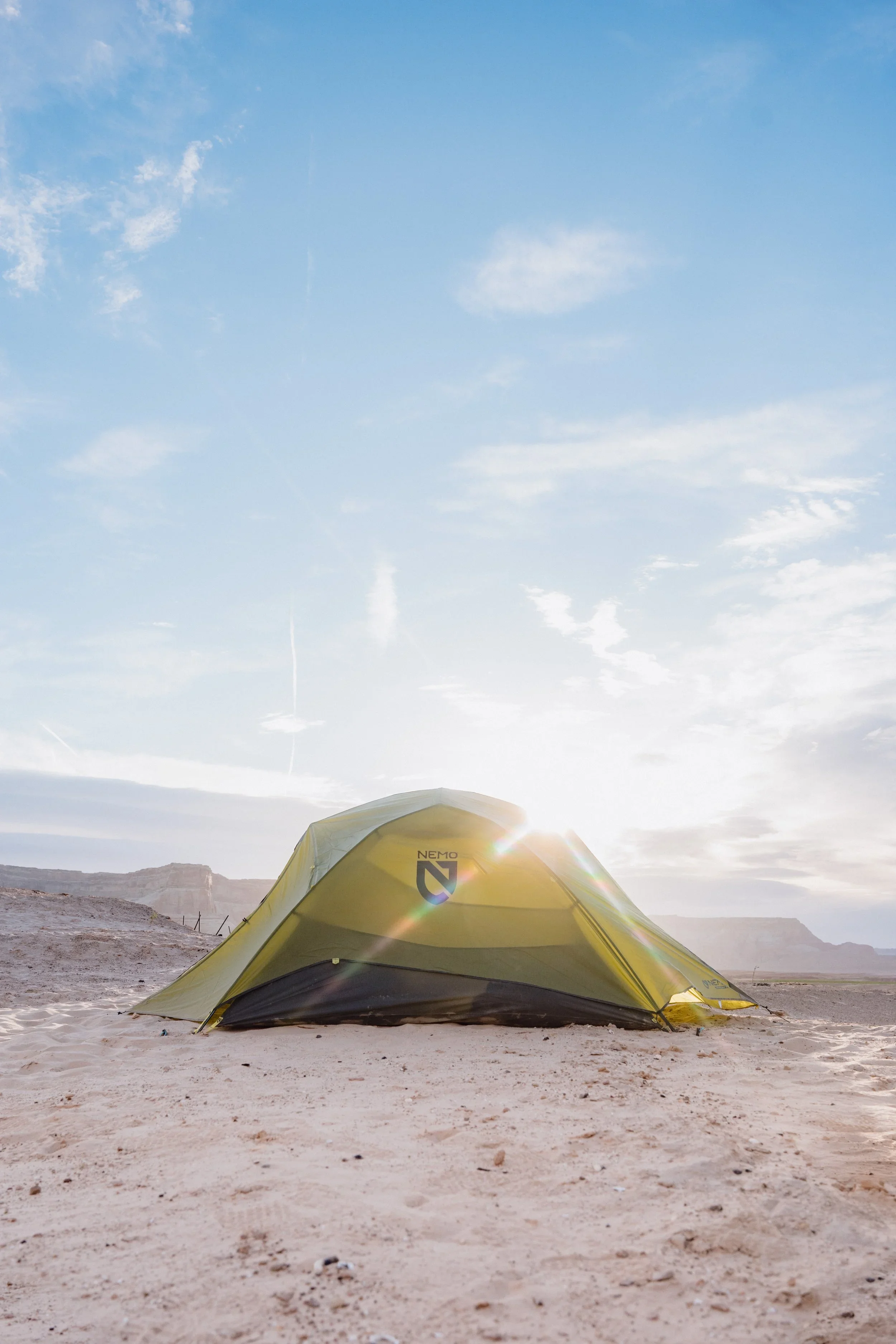 A yellow tent set up on sandy desert ground with mountains in the background, under a partly cloudy sky at sunrise or sunset.