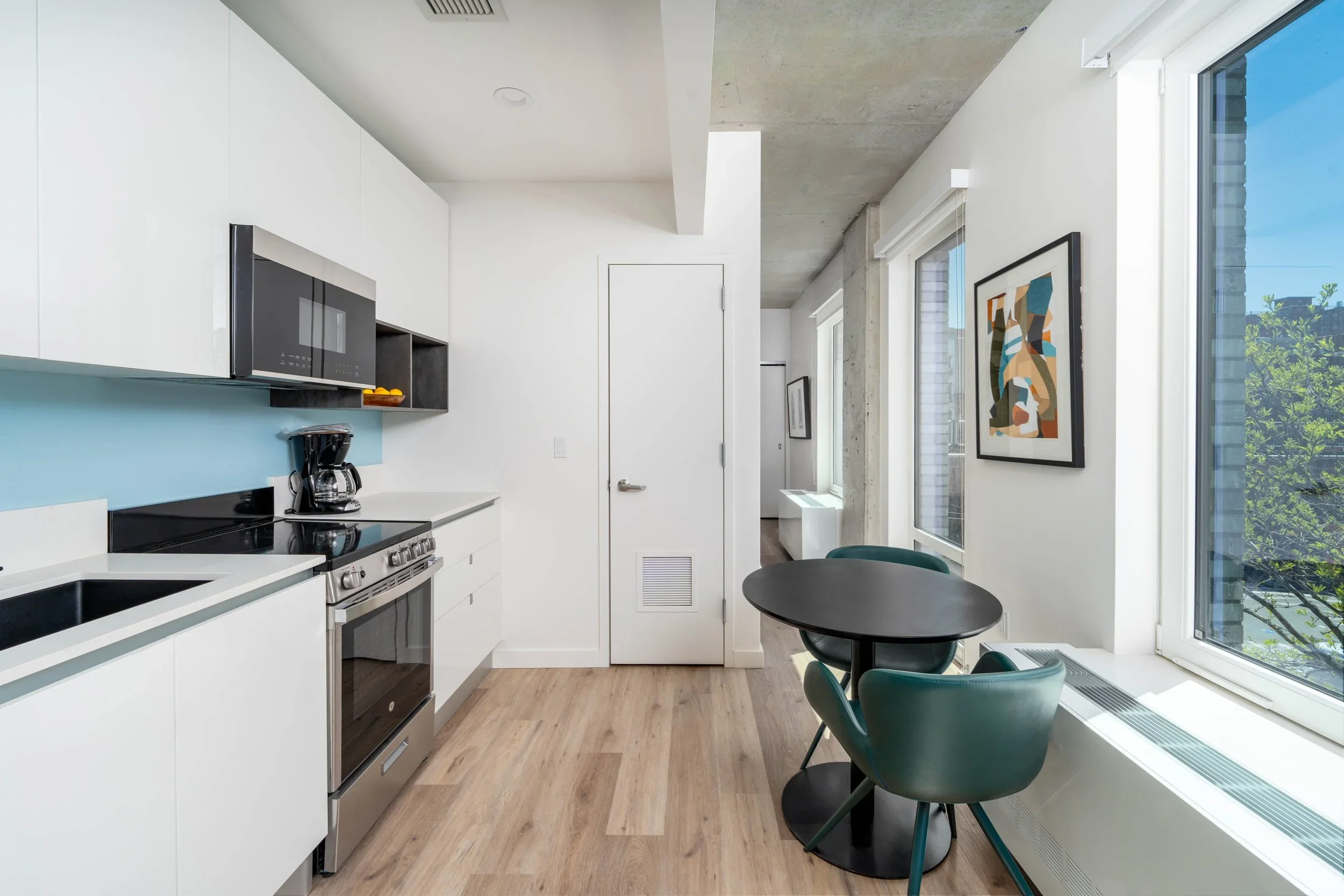 Modern kitchen and dining area with white cabinets, a black stove, microwave, a round black dining table, and green chairs, large windows showing trees outside, with minimalistic decorations.