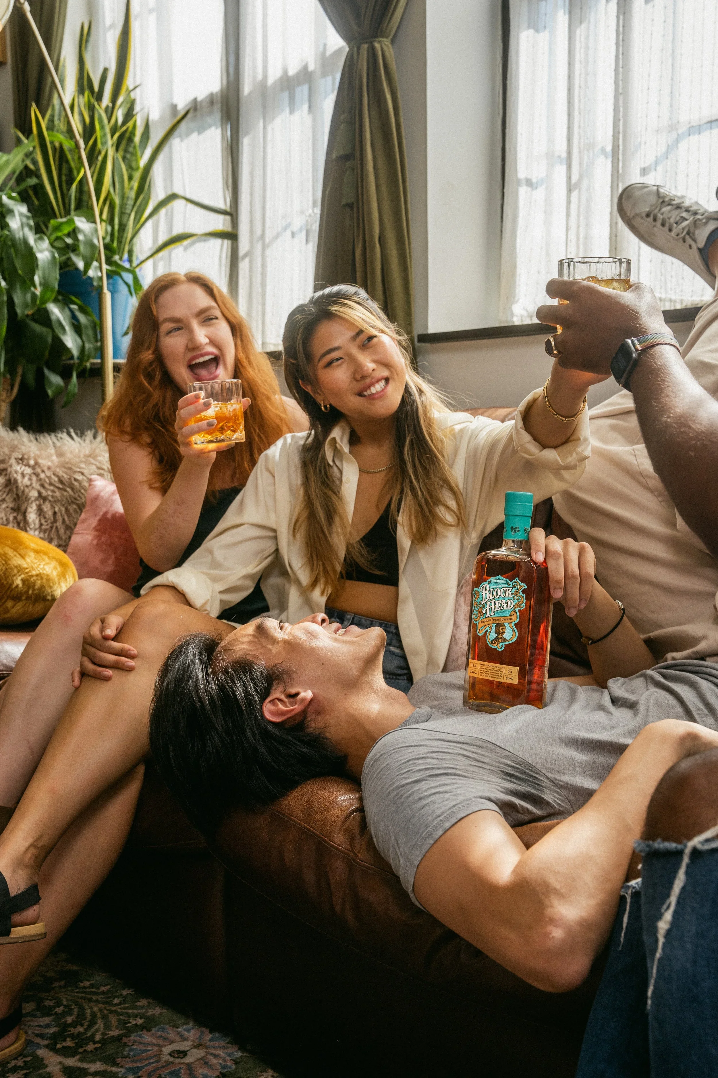 Four friends relaxing and having fun indoors; one lying on a couch with a bottle of liquor, two sitting behind, one with a drink, and one taking a selfie.