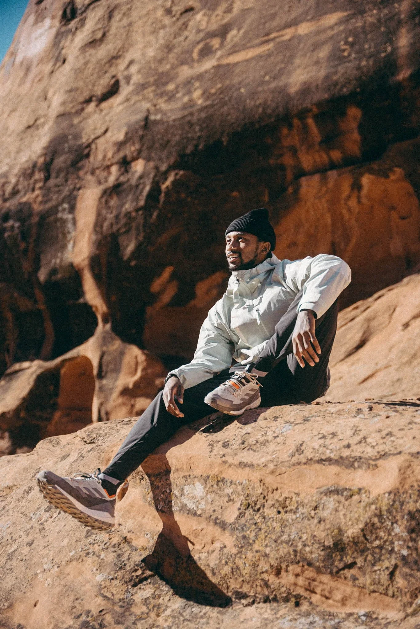 A man sitting on a large rock in a desert landscape, wearing a black beanie, light jacket, dark pants, and Adidas sneakers, with a rocky cliff in the background.