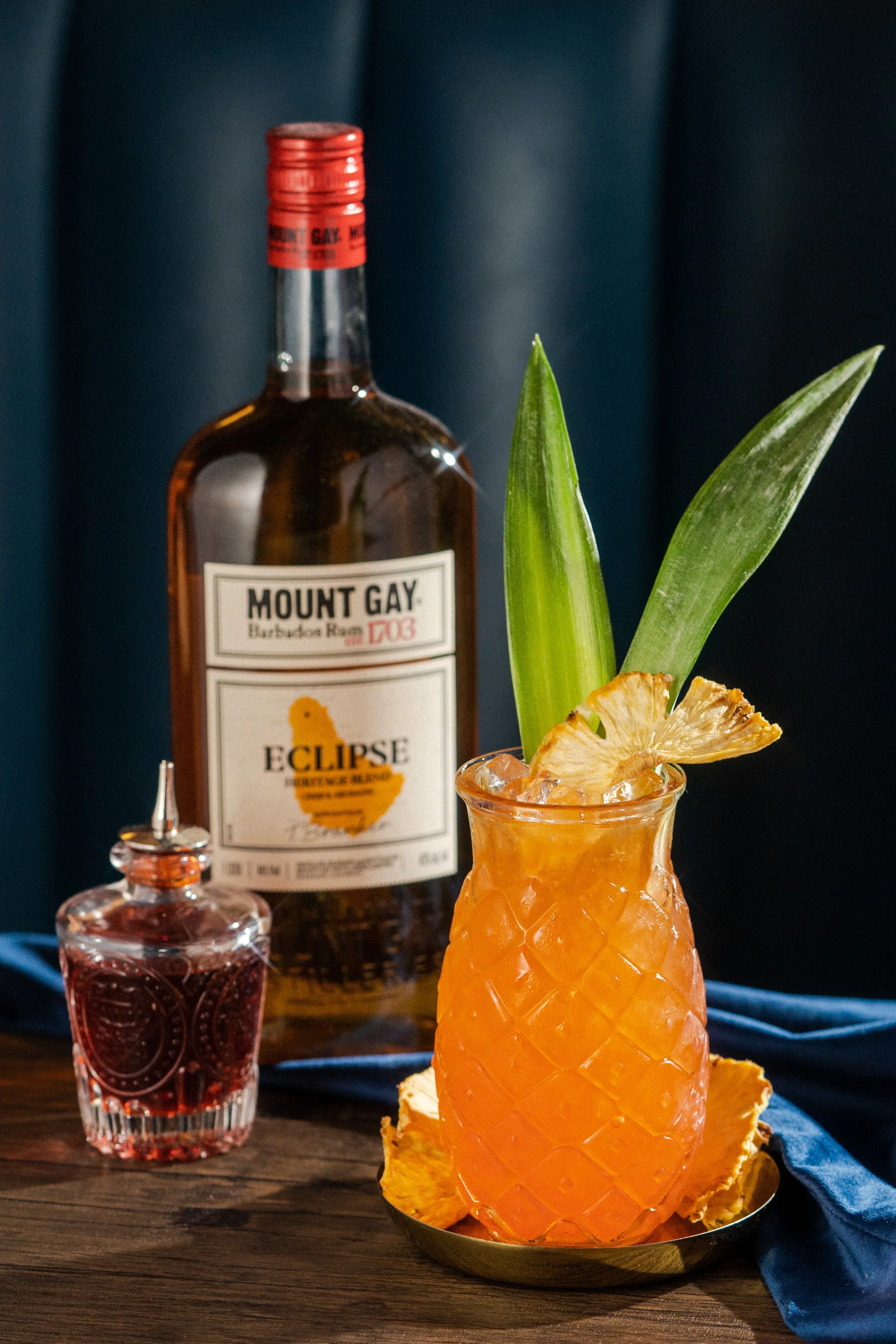 A tropical cocktail served in a pineapple-shaped glass, garnished with pineapple slices and green leaves, with a bottle of Mount Gay Eclipse rum and a small bottle of grenadine nearby on a wooden surface.