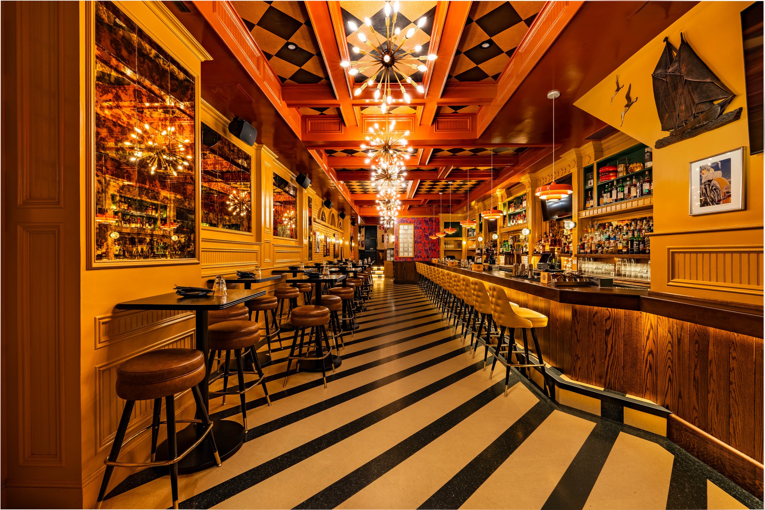 Interior of a stylish bar with a long counter, rows of bar stools, warm lighting, and decorative wall art.