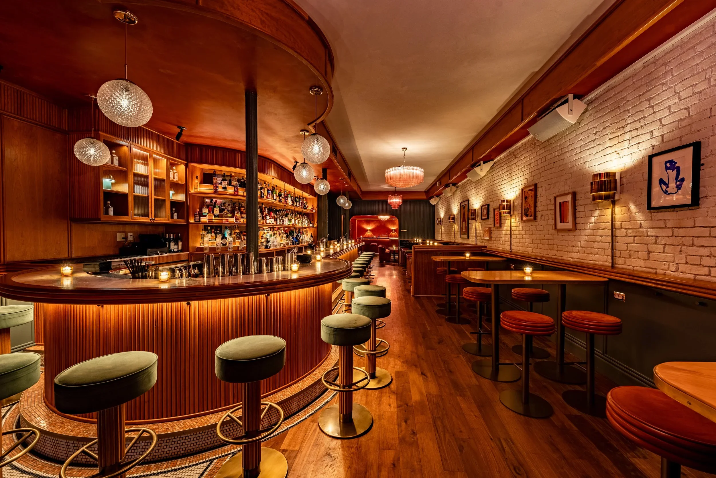 Empty bar with wooden counter, green and brown stools, paintings on white brick wall, dotted lighting fixtures, and a cozy atmosphere.