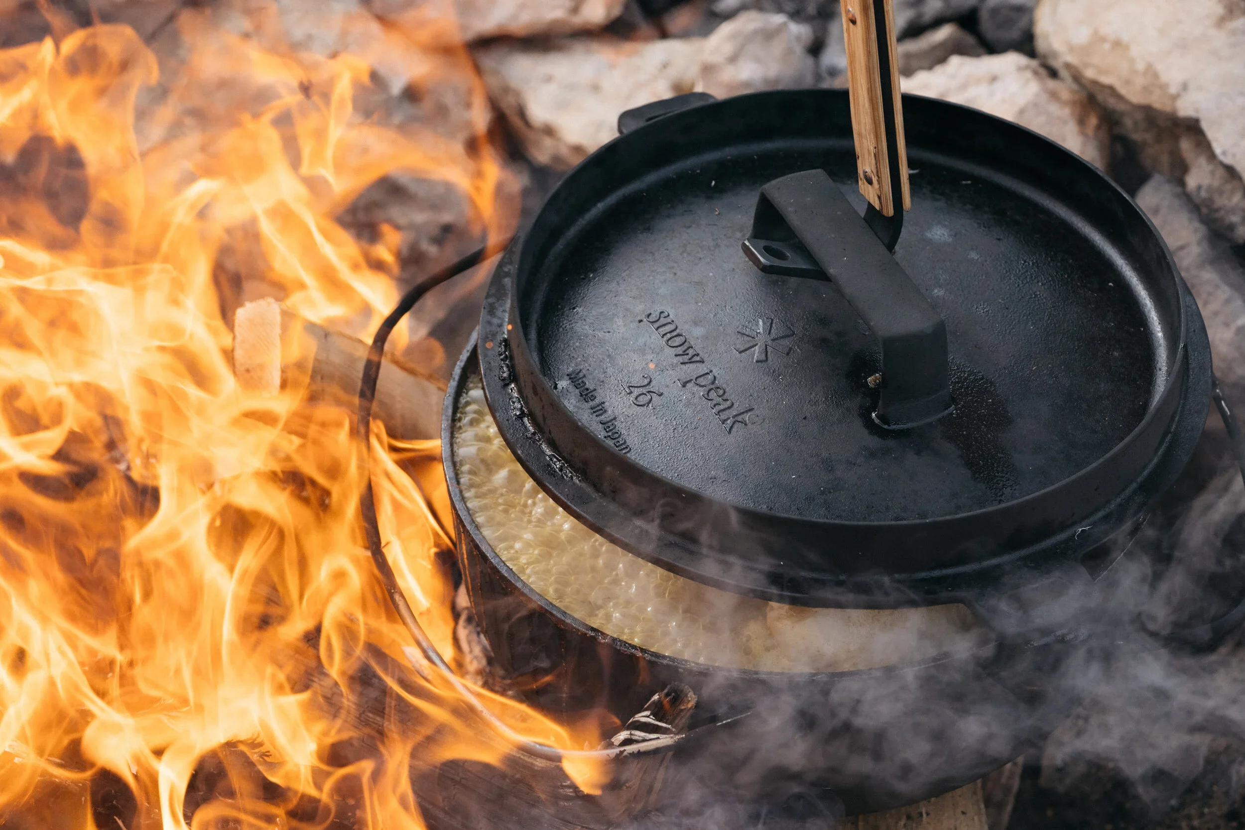 A cast iron Dutch oven with a lid over a fire outdoors, likely cooking food.