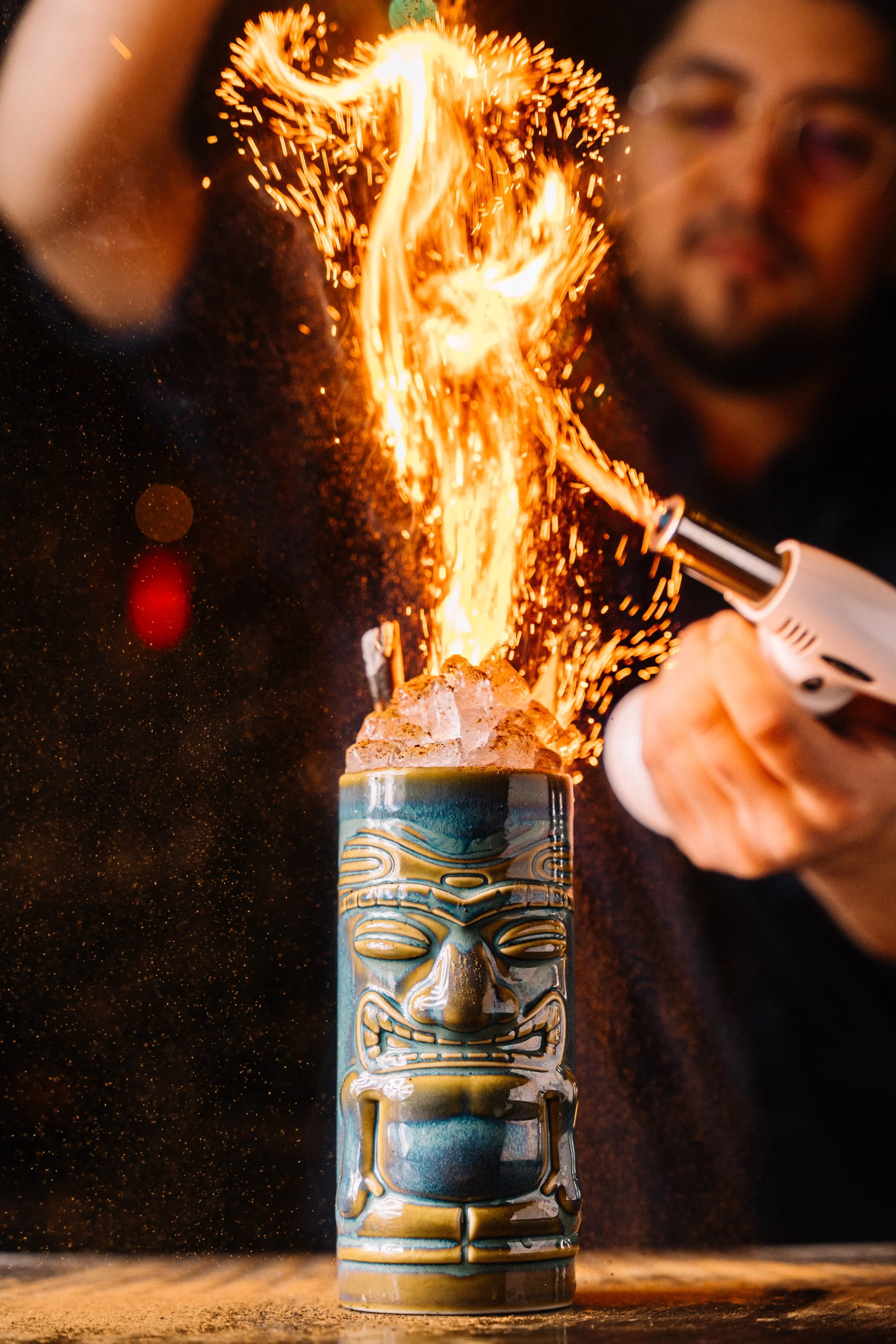 A person holding a torch igniting a tiki-style mug with flames and sparks, with a blurred background.