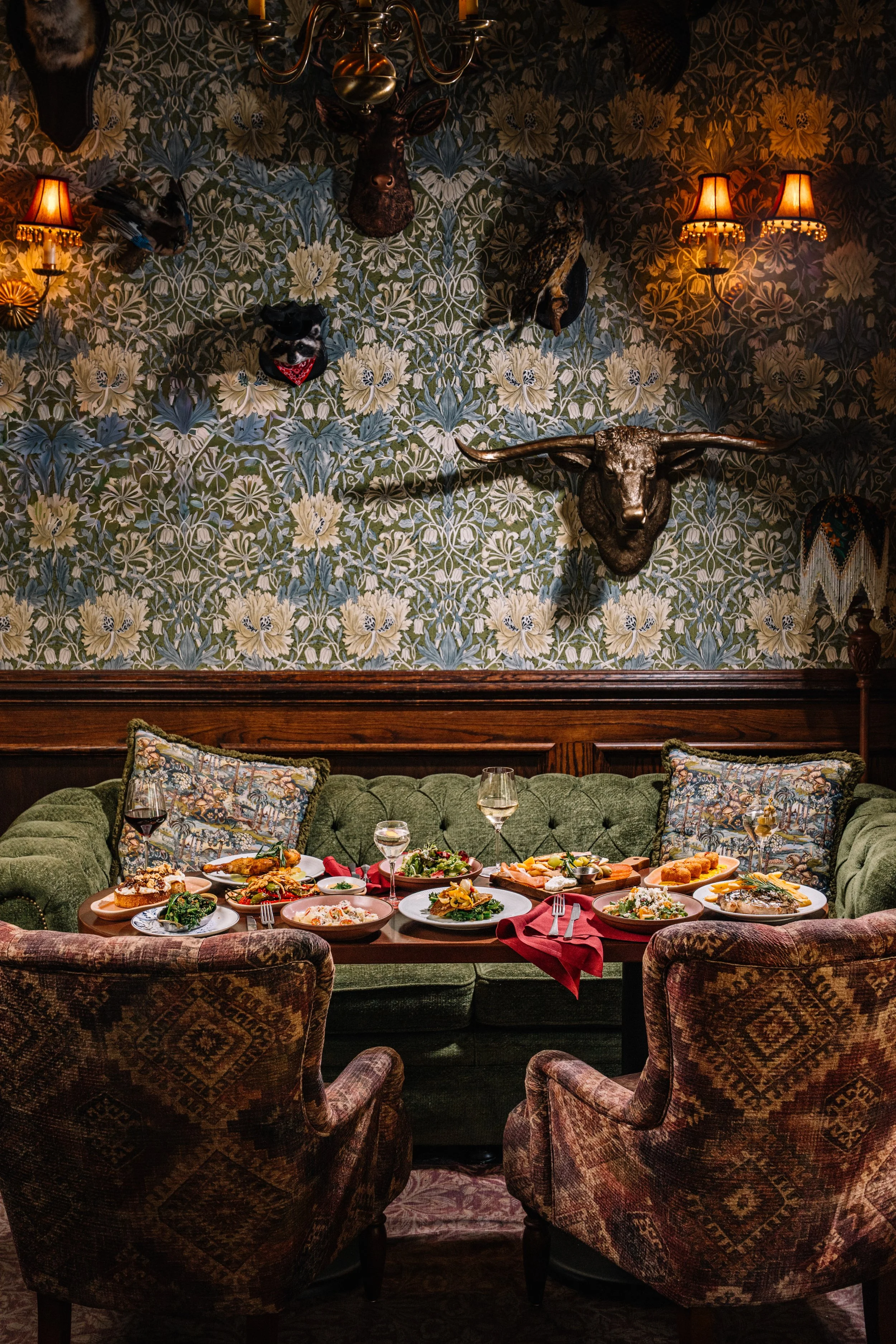 A dining room with a green velvet sofa and upholstered armchairs, set with plates of food, wine glasses, and cutlery. The wall behind features floral wallpaper, mounted animal heads, including a buffalo skull with horns, a bear head, and a bird, with
