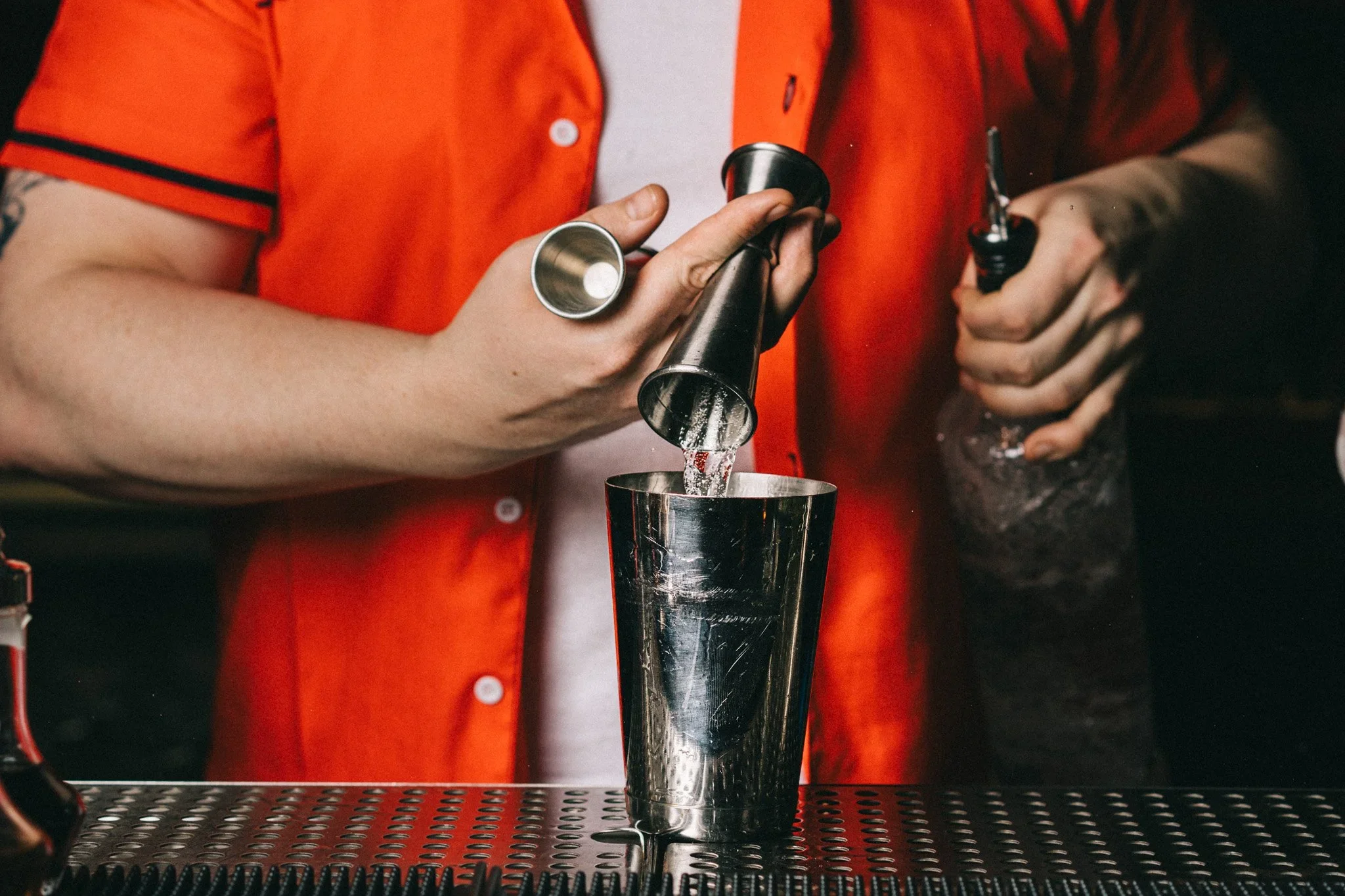 A bartender in an orange shirt pouring liquid from a jigger into a cocktail shaker.