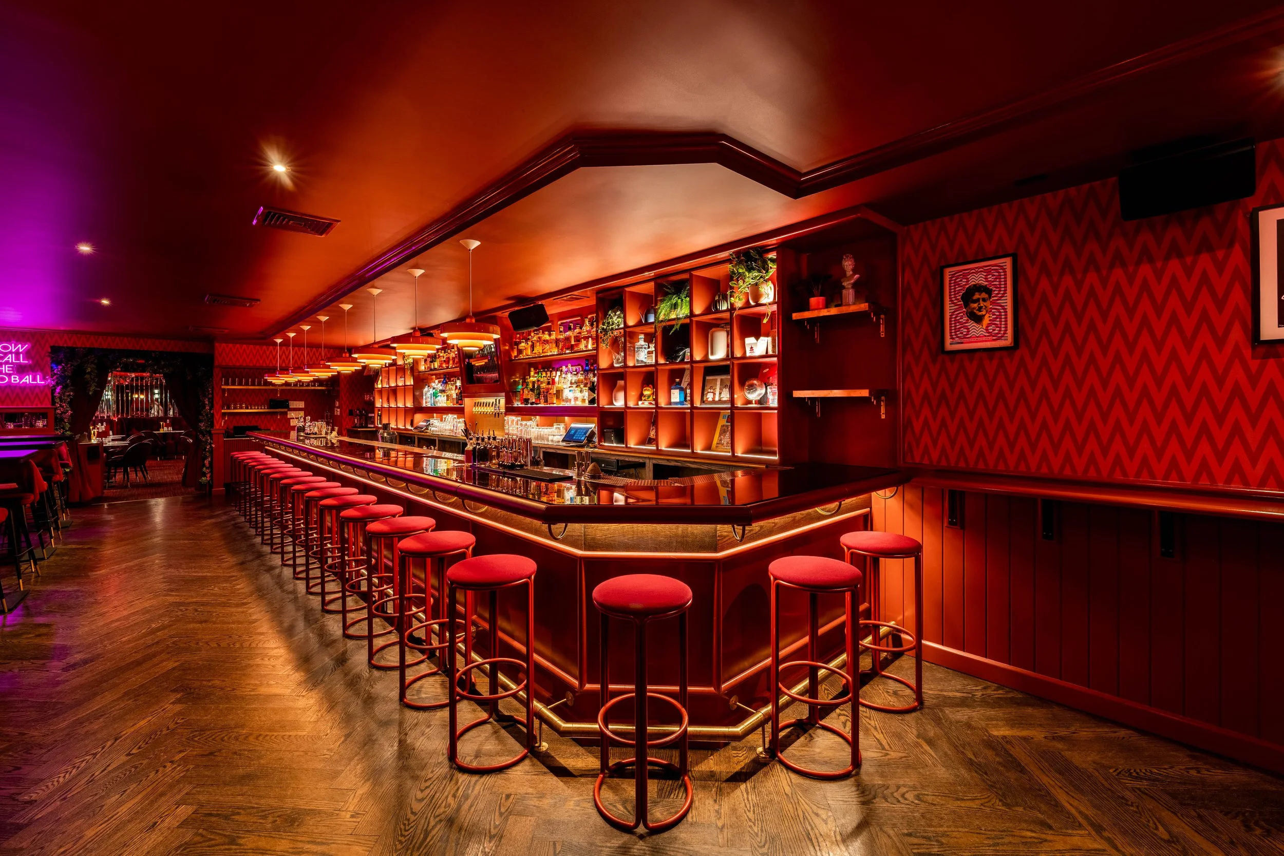 A dimly lit bar with red decor, a long curved counter, and red bar stools. Shelves behind the bar display various bottles and decorative items, with some greenery. Dark wood flooring and modern lighting create a cozy atmosphere.