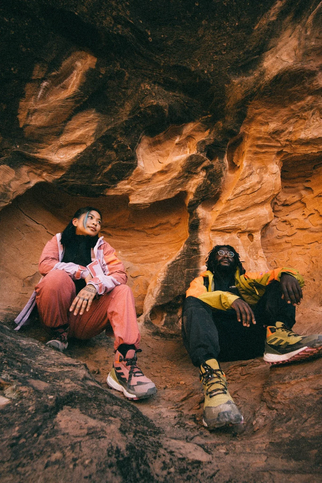 Two people sitting inside a red rock canyon, wearing outdoor hiking gear and shoes.