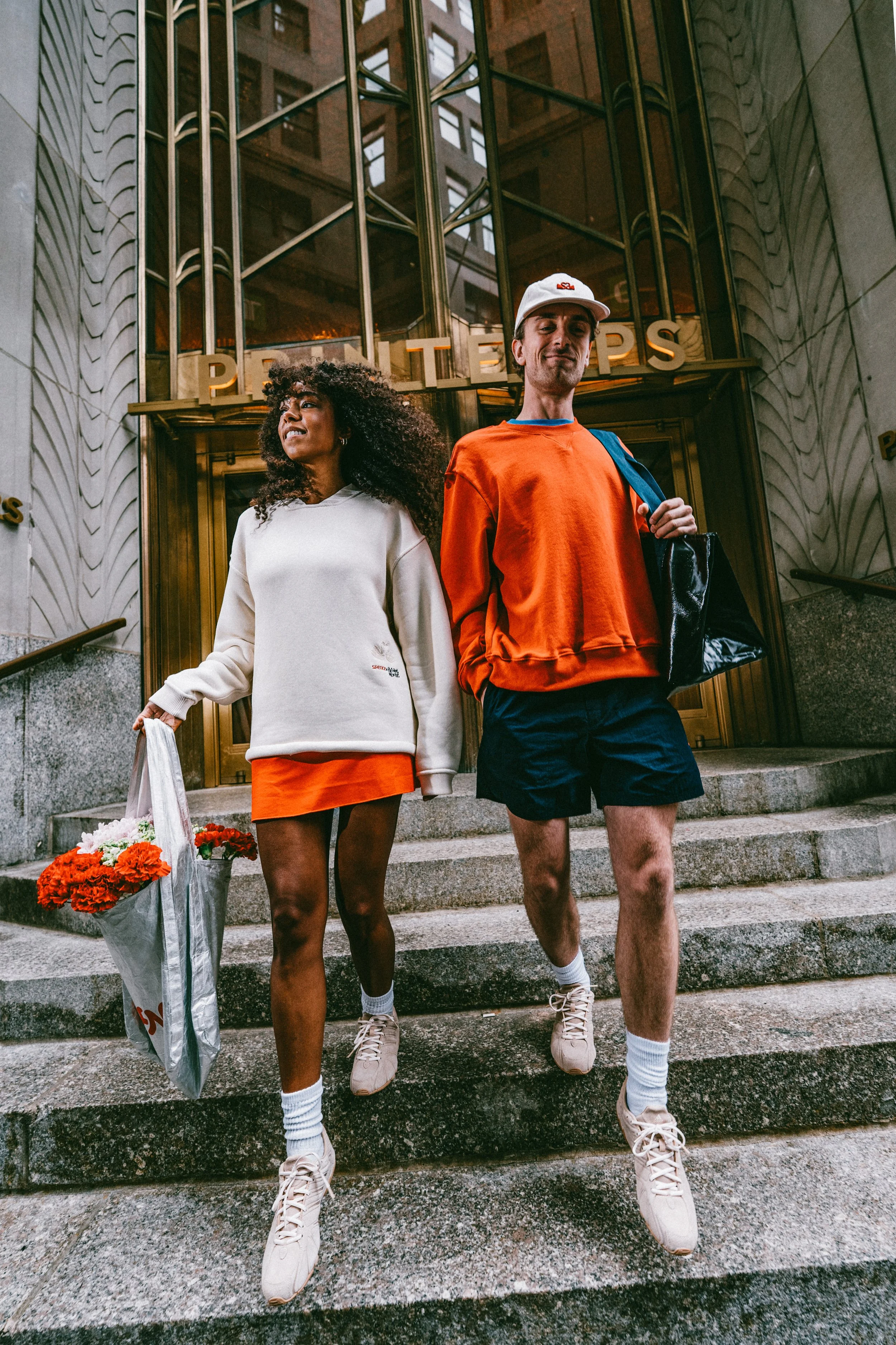 Two young adults, a woman with curly hair holding a bag of flowers and a man with a cap carrying a bag, walking down the steps outside a building with 