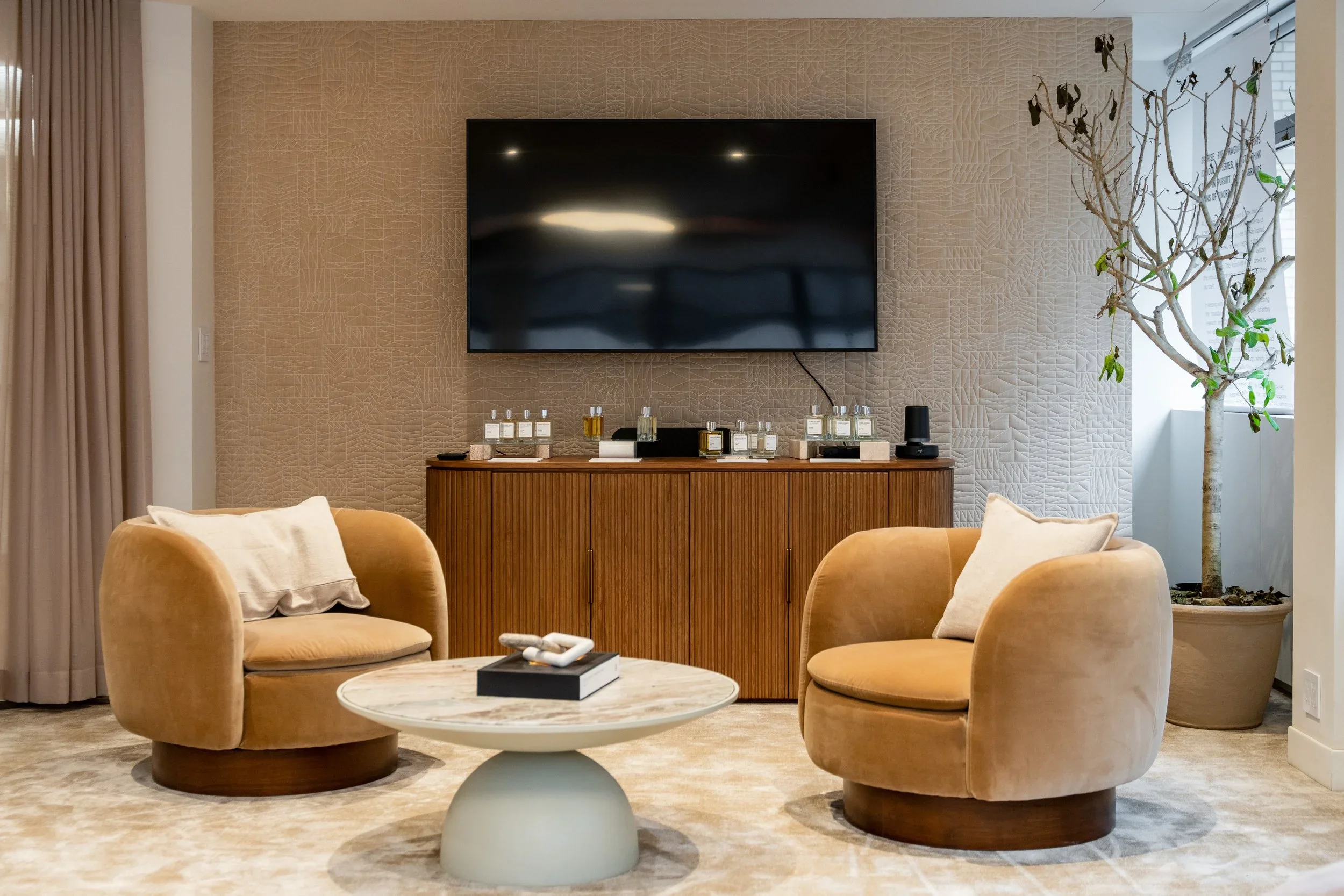 A modern living room with two beige rounded armchairs, a round marble coffee table, a textured beige wall with a mounted flat-screen TV, a wooden cabinet with perfume bottles and decorative items, and a potted tree near a window with beige curtains.