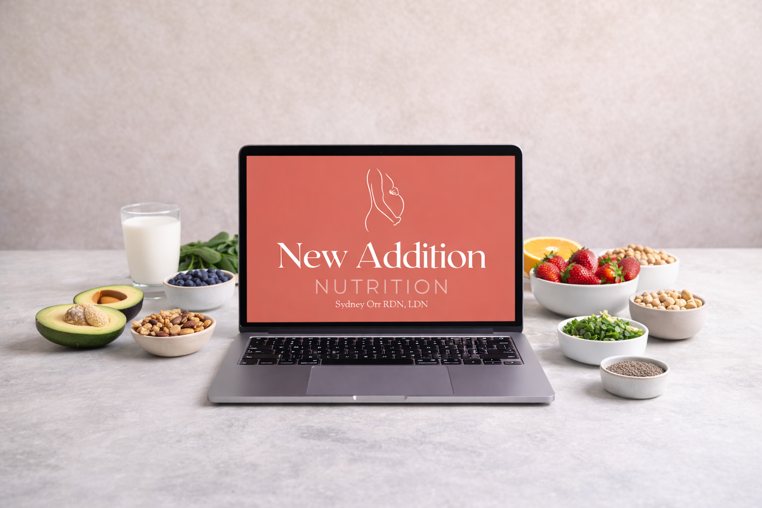 Laptop displaying 'New Addition Nutrition' with berries, avocados, nuts, seeds, lemon, and a glass of milk surrounding it on a grey surface.