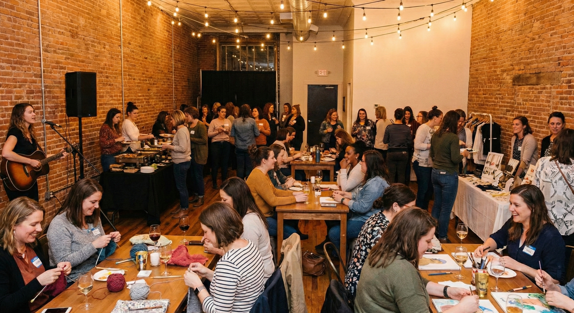 Women at a social gathering or workshop in a warmly lit brick-walled room, some sitting at tables with drinks and knitting, others browsing merchandise or chatting, with a musician playing guitar on the left.