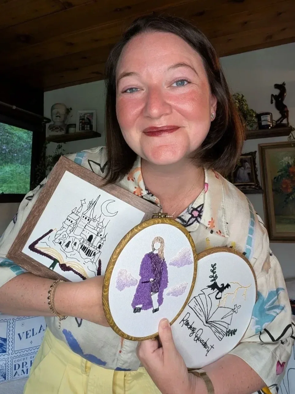 A woman with short brown hair and blue eyes smiling while holding three embroidered artworks, including a castle, Taylor Swift in a purple dress, and a witch flying on a broomstick, in a room with wooden ceilings and shelves.