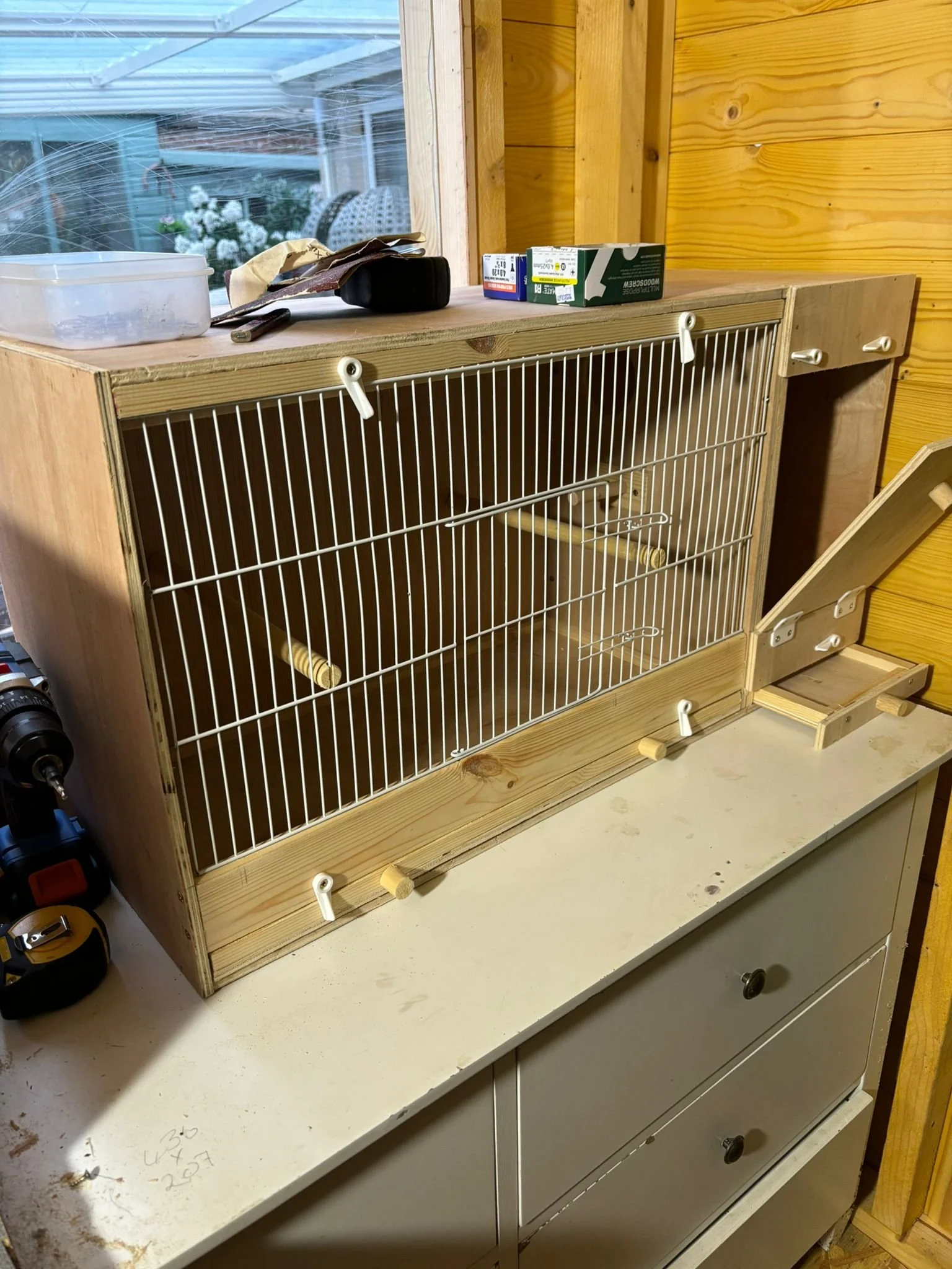 Unfinished wooden and metal birdcage with a slide-out tray, placed on a white cabinet in a workshop or shed. On top of the cage, there are tools and some boxes, with a window and outside scenery in the background.