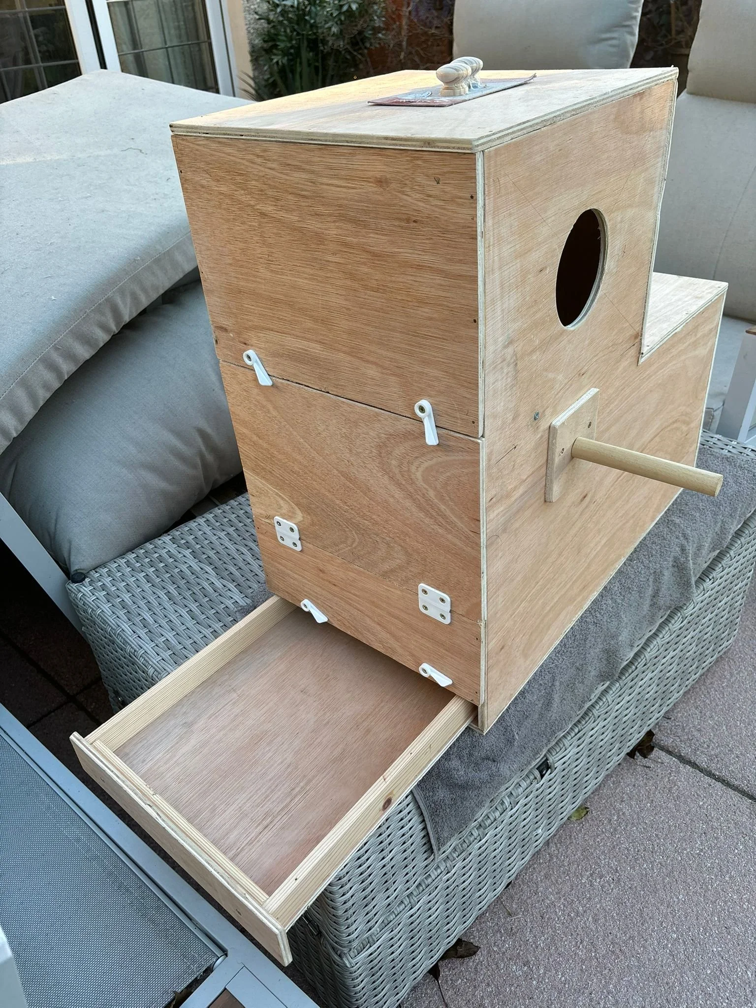 Unfinished wooden birdhouse with a sliding drawer at the bottom, a circular entry hole on the side, and a perch outside the hole, placed on a patio sofa.