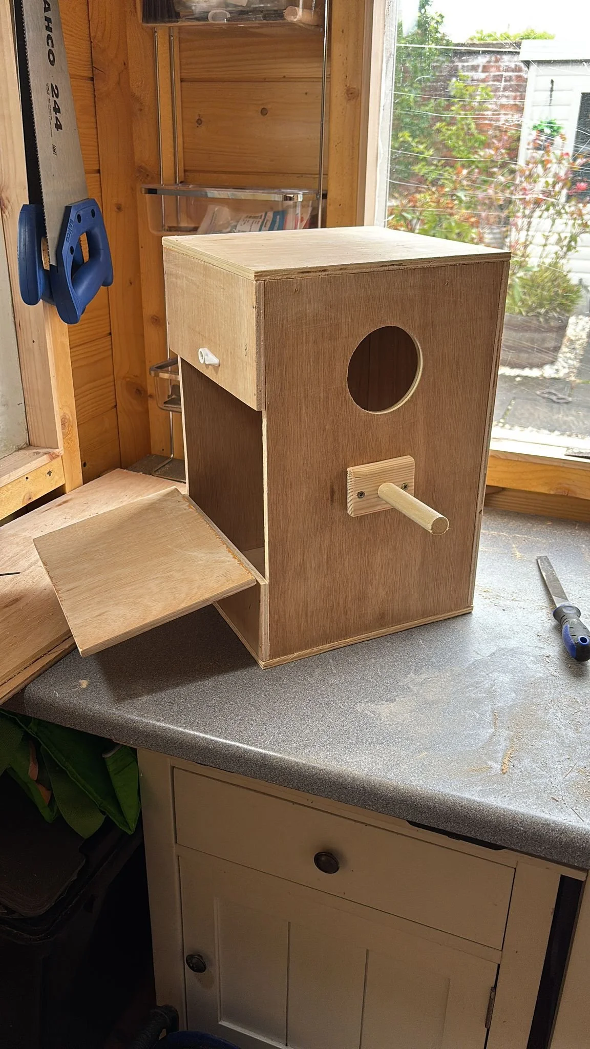 A partially assembled wooden birdhouse on a workbench in a workshop, with a circular entry hole, a perch, and a small roof overhang, near a window with a garden view.