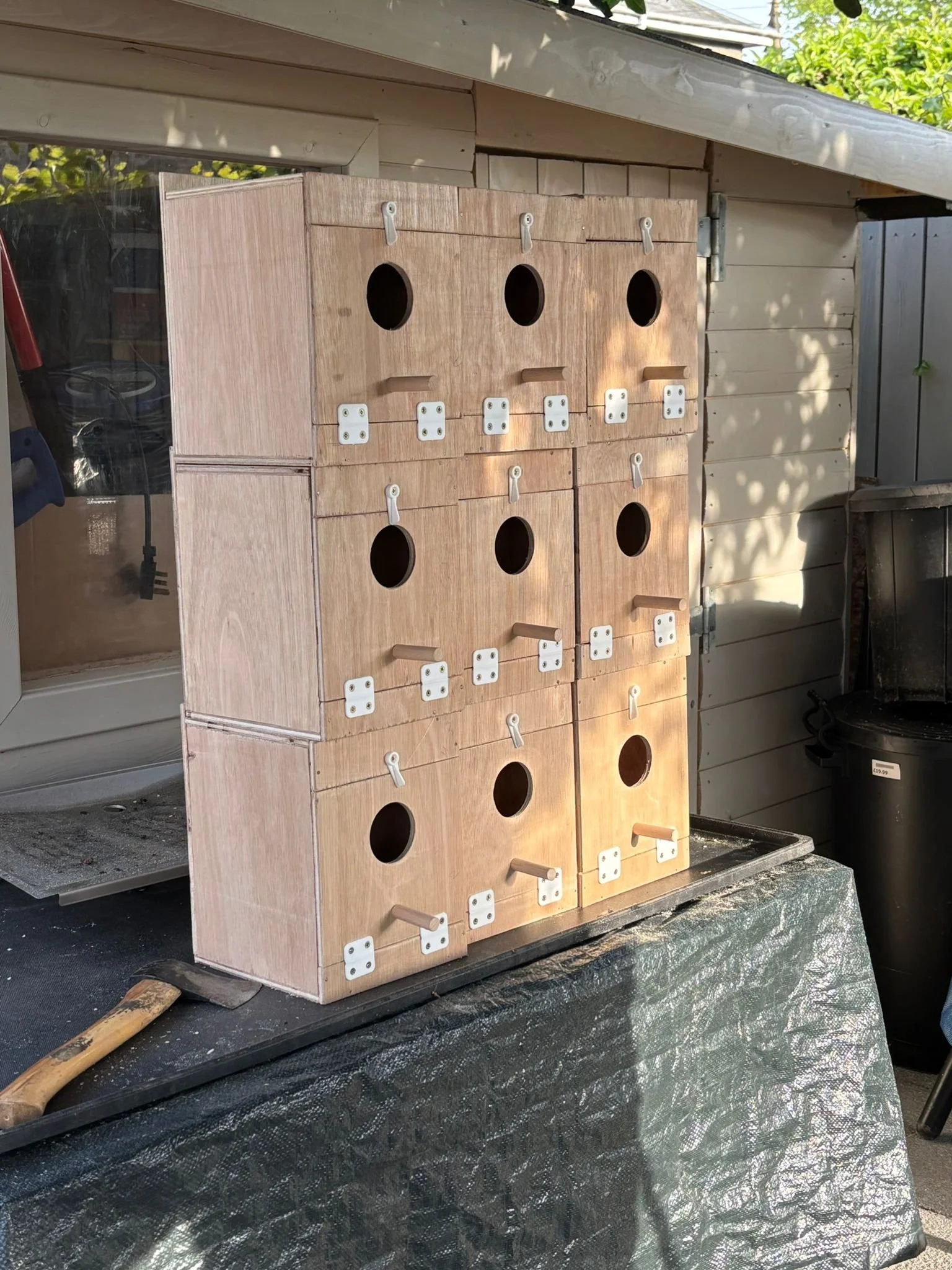 Stacked wooden birdhouses with small round entry holes, hooks, and latch mechanisms, sitting on a table outdoors.
