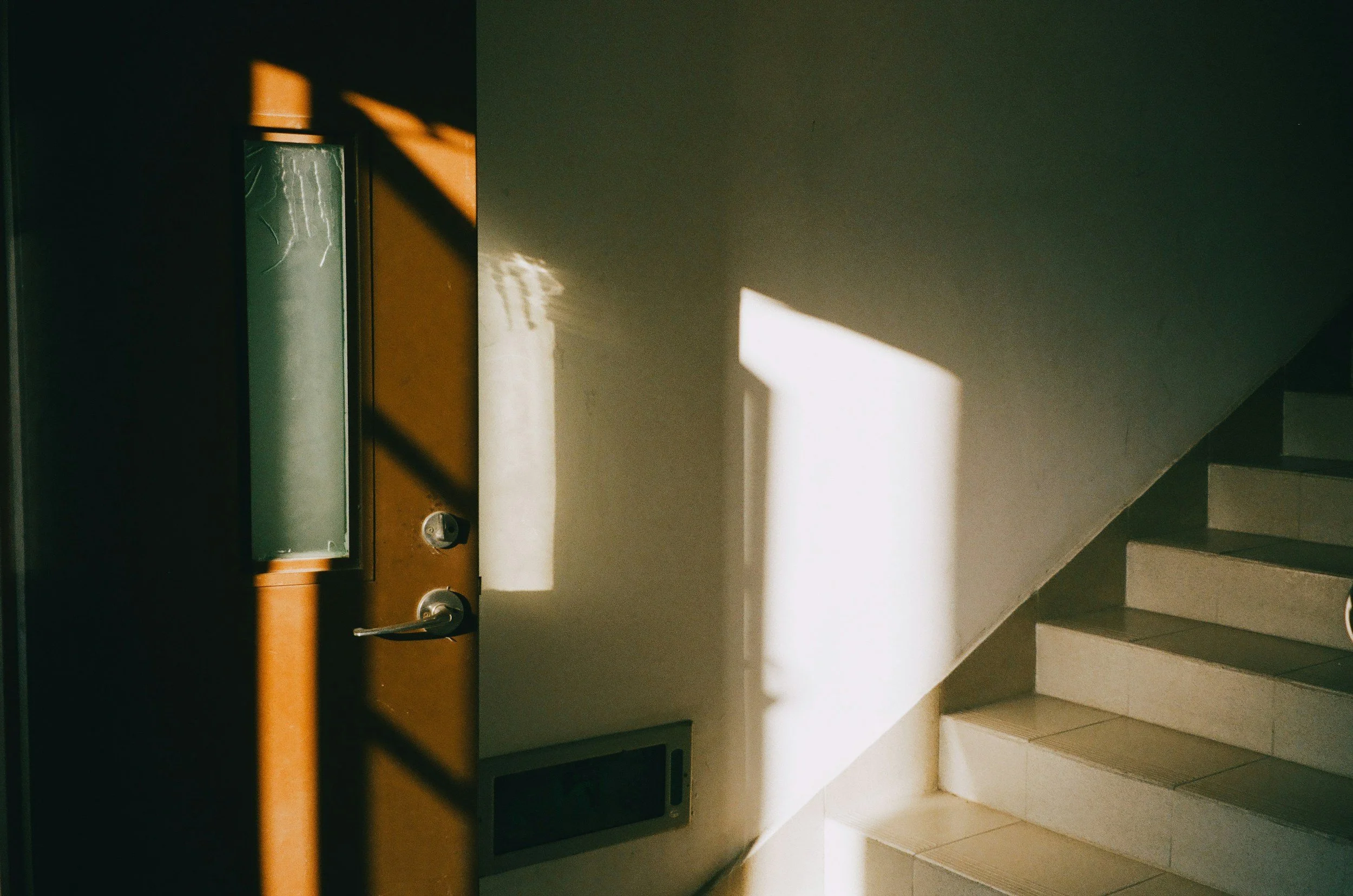 Sunlight casting a shadow of a window frame on a white wall next to a staircase with tiled steps.