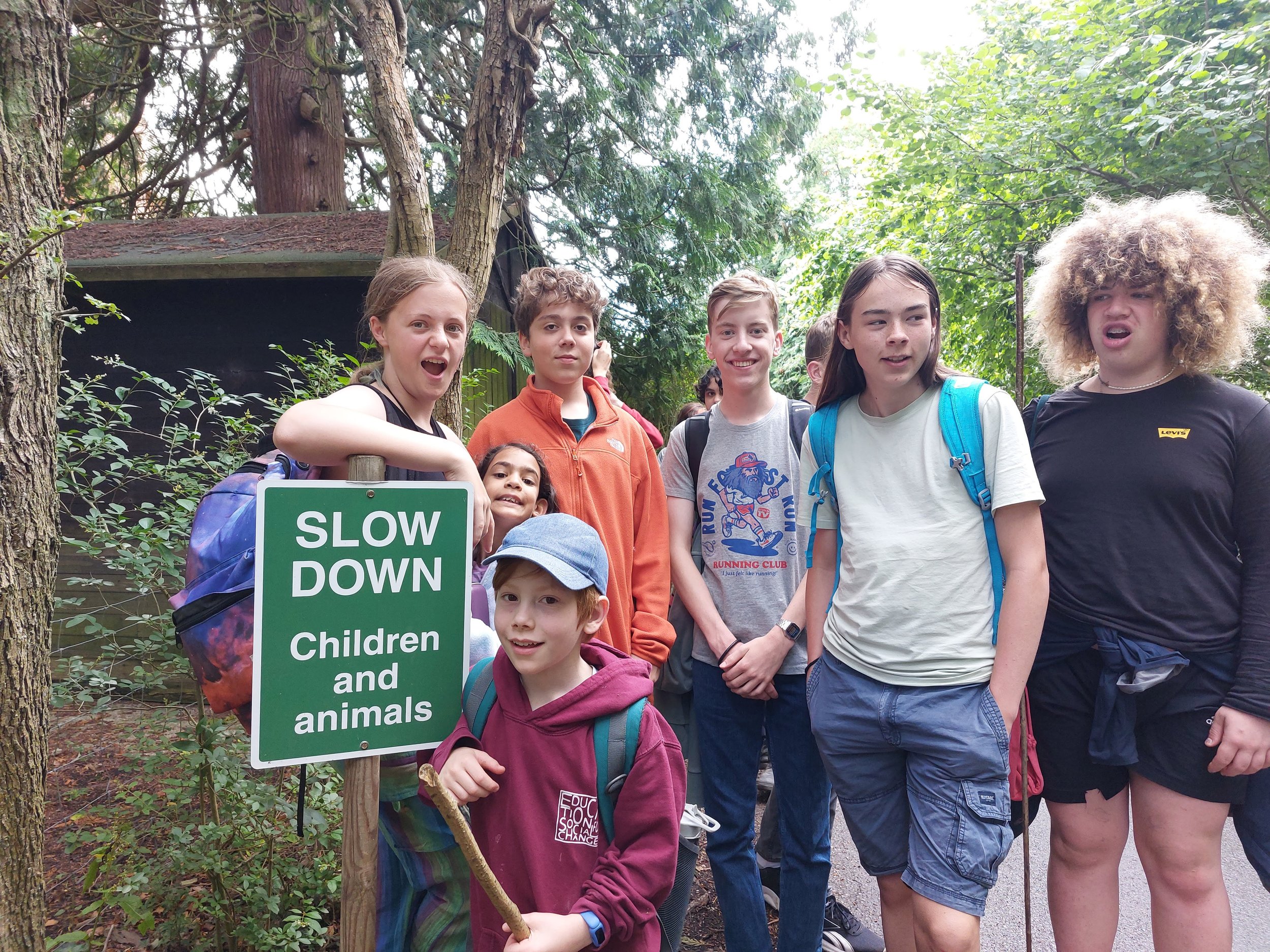 Group of children and teenagers standing outdoors in a forested area near a green sign that reads 'SLOW DOWN Children and animals'.