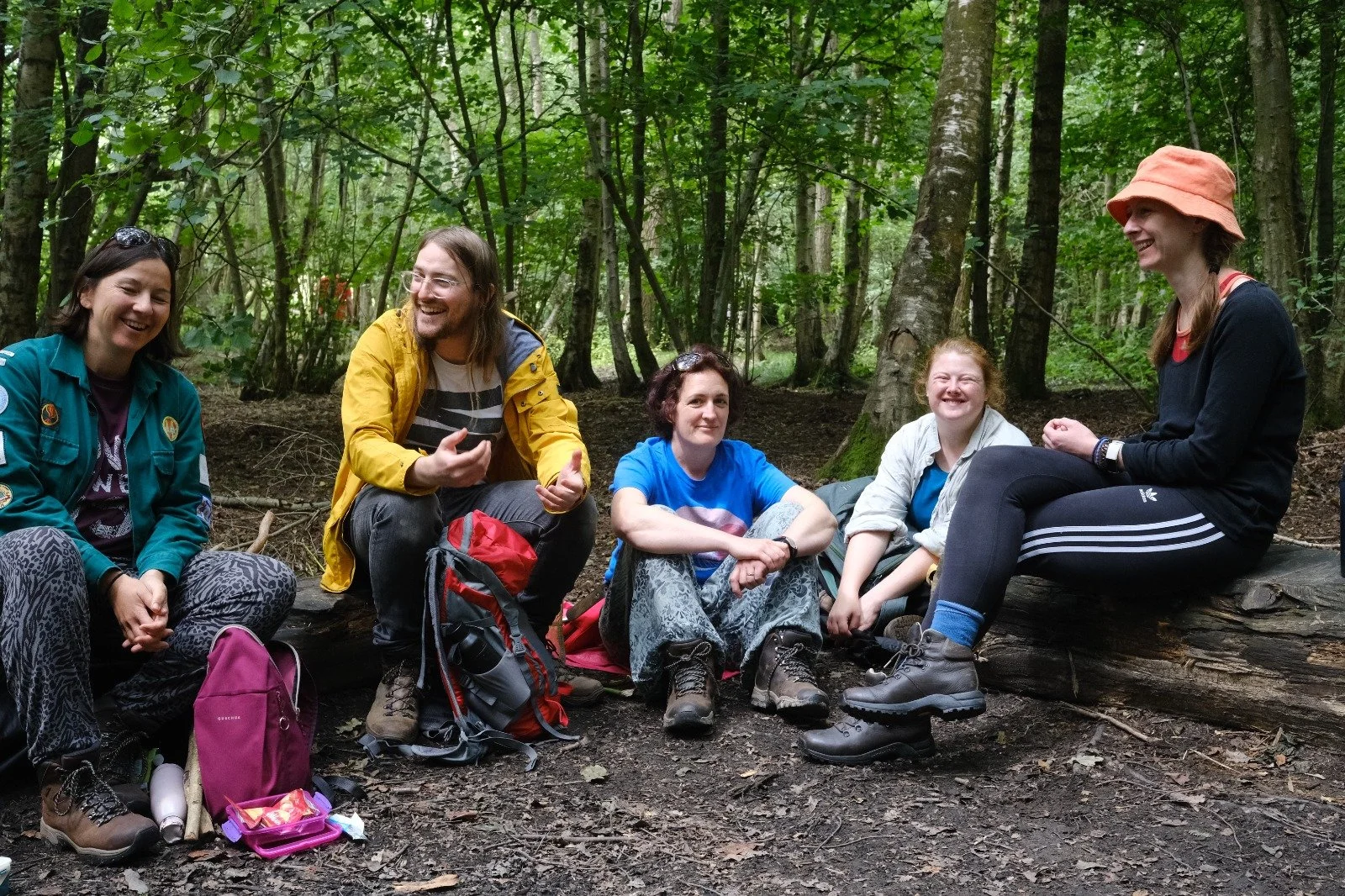 A group of six friends sitting on logs in a forest, smiling and enjoying each other's company. They are wearing outdoor clothing, with backpacks and hiking gear.
