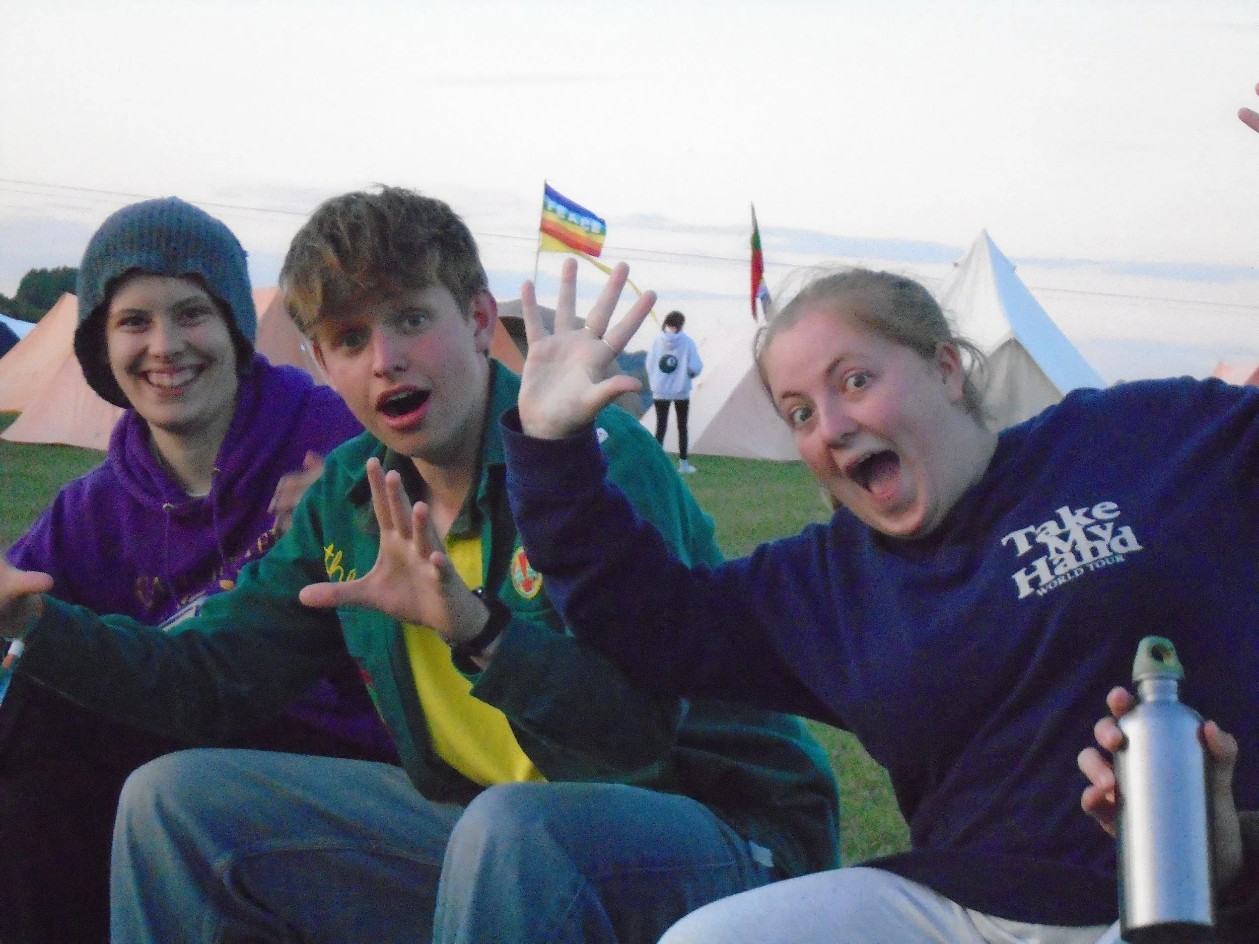 Three young people smiling and posing for a selfie at an outdoor event with tents and flags in the background.