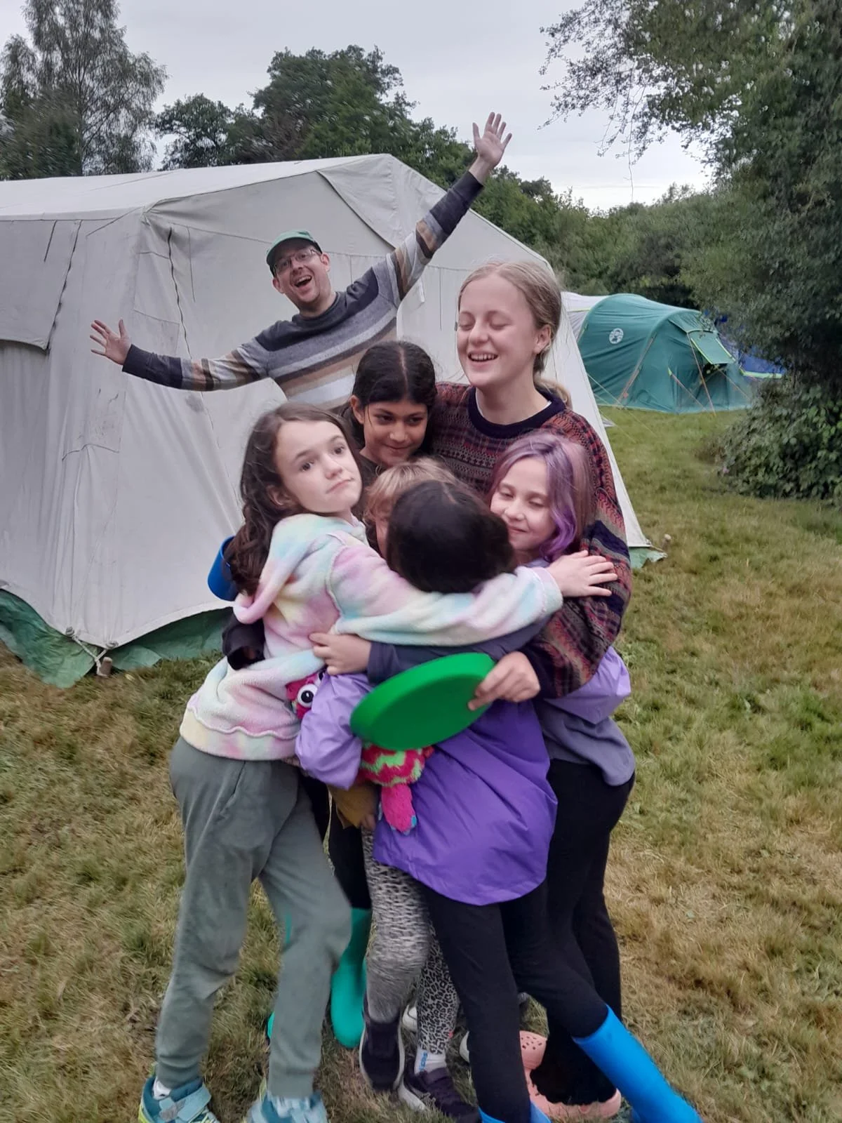 A group of six children hugging each other in front of a large camping tent outdoors, with two more tents visible in the background and trees surrounding the area.