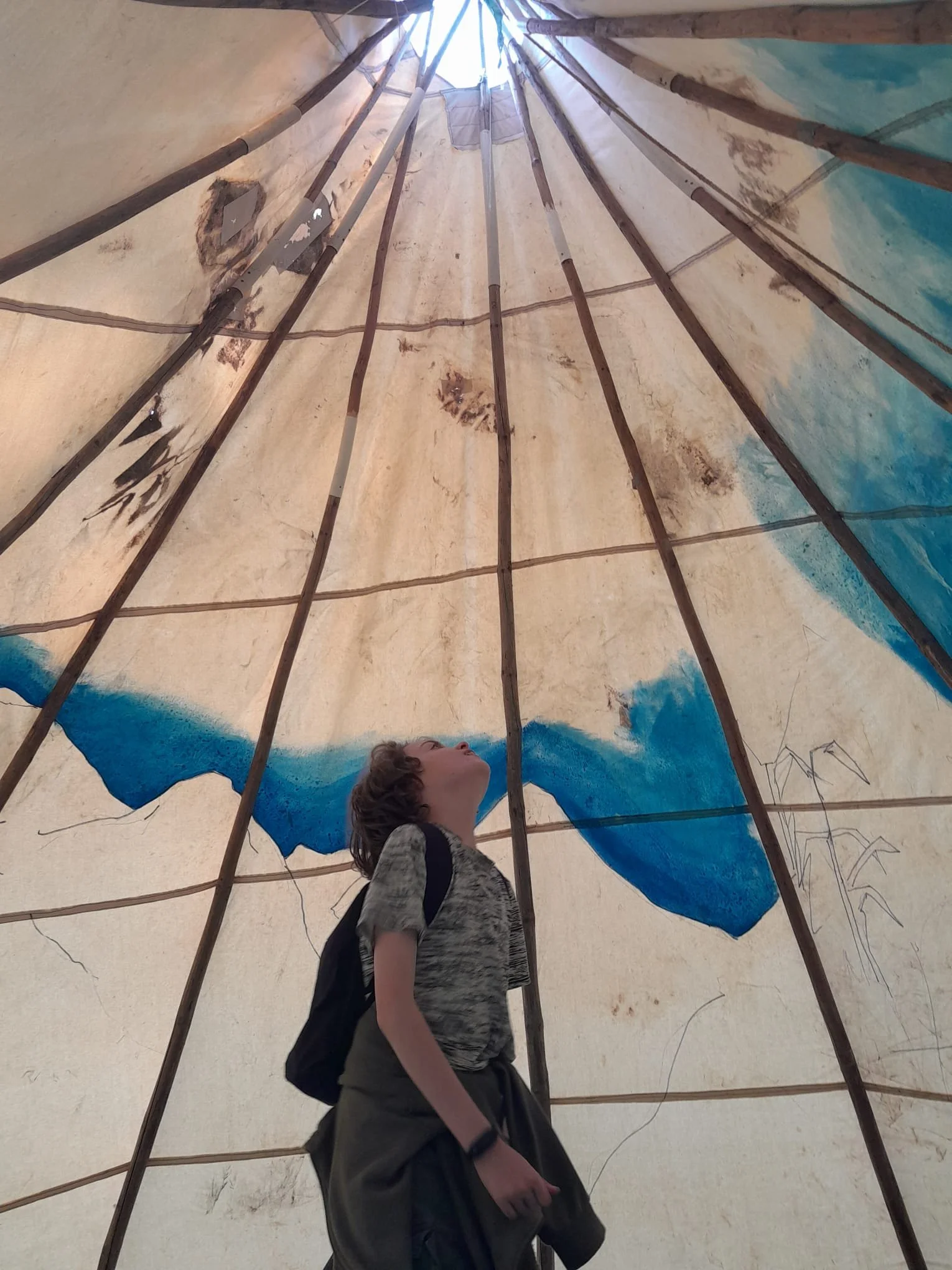 A person standing inside a tipi or teepee with a painted design on the fabric, looking upward towards the top opening, with wooden poles supporting the structure.