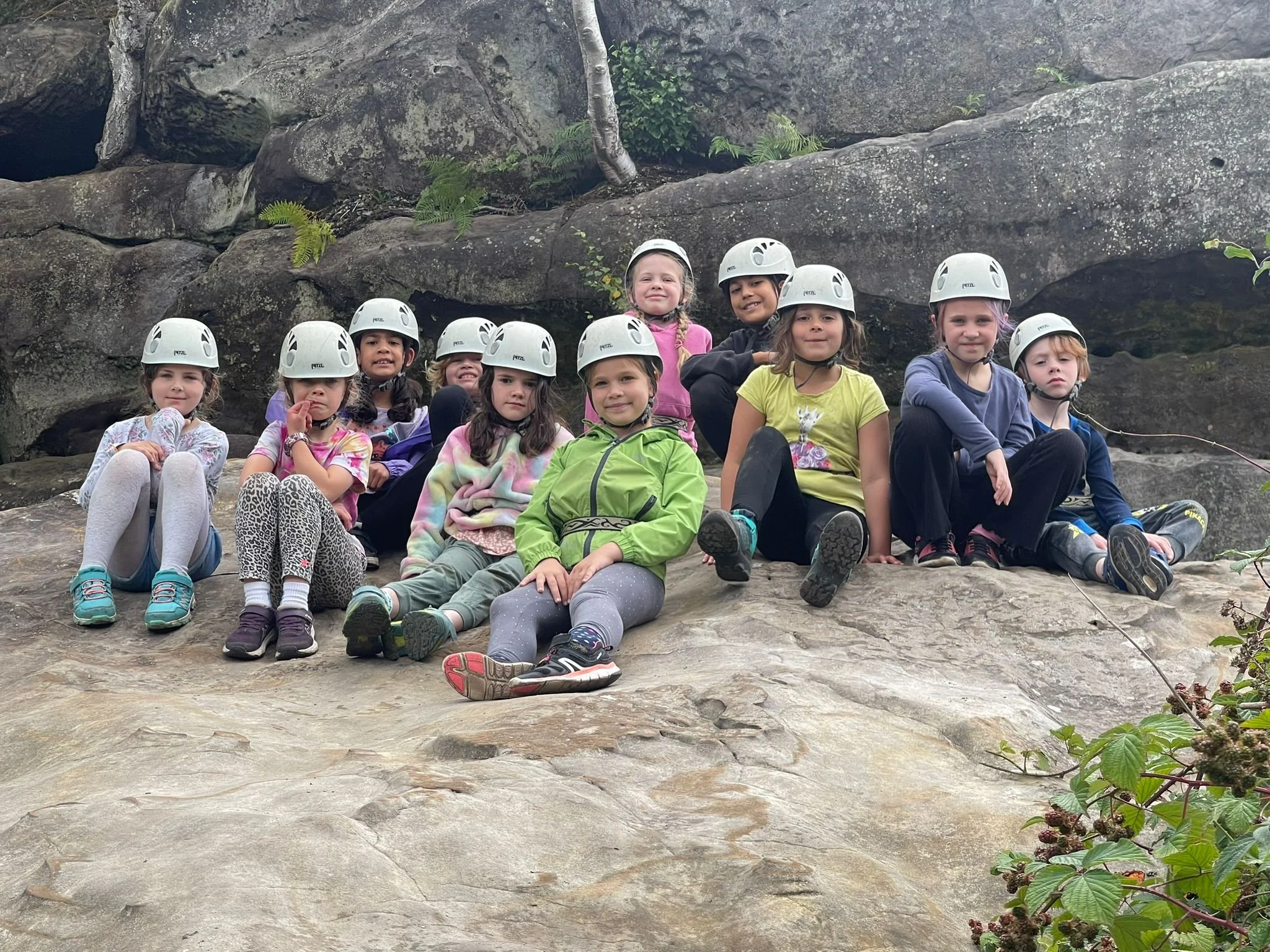A group of children wearing white helmets and casual clothing, sitting on a large flat rock surface in front of a rocky landscape with some green plants, smiling and looking at the camera.