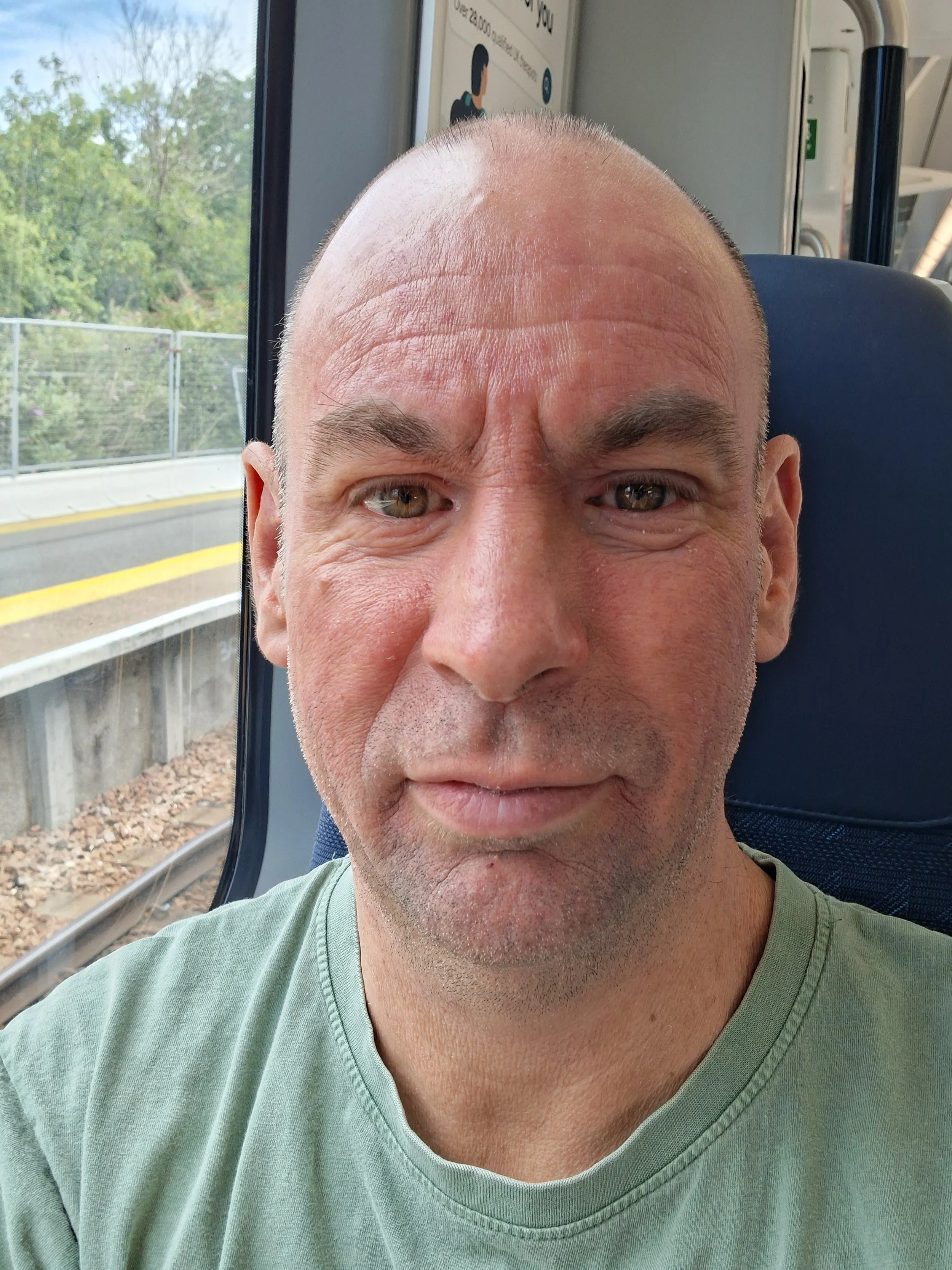 A close-up photo of a smiling middle-aged man with a shaved head and green eyes, wearing a light green t-shirt, sitting on a bus or train with a window showing trees and a fence outside.
