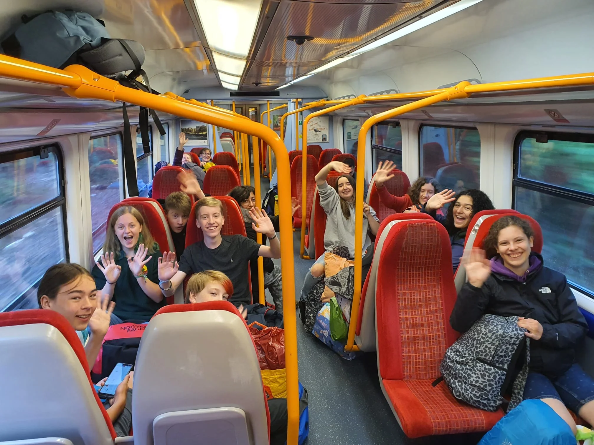 The inside of a bus filled with smiling children and teenagers waving at the camera. The bus has red upholstered seats and yellow handrails, and there are backpacks and belongings near the seats. The windows show a blurred outdoor scene.