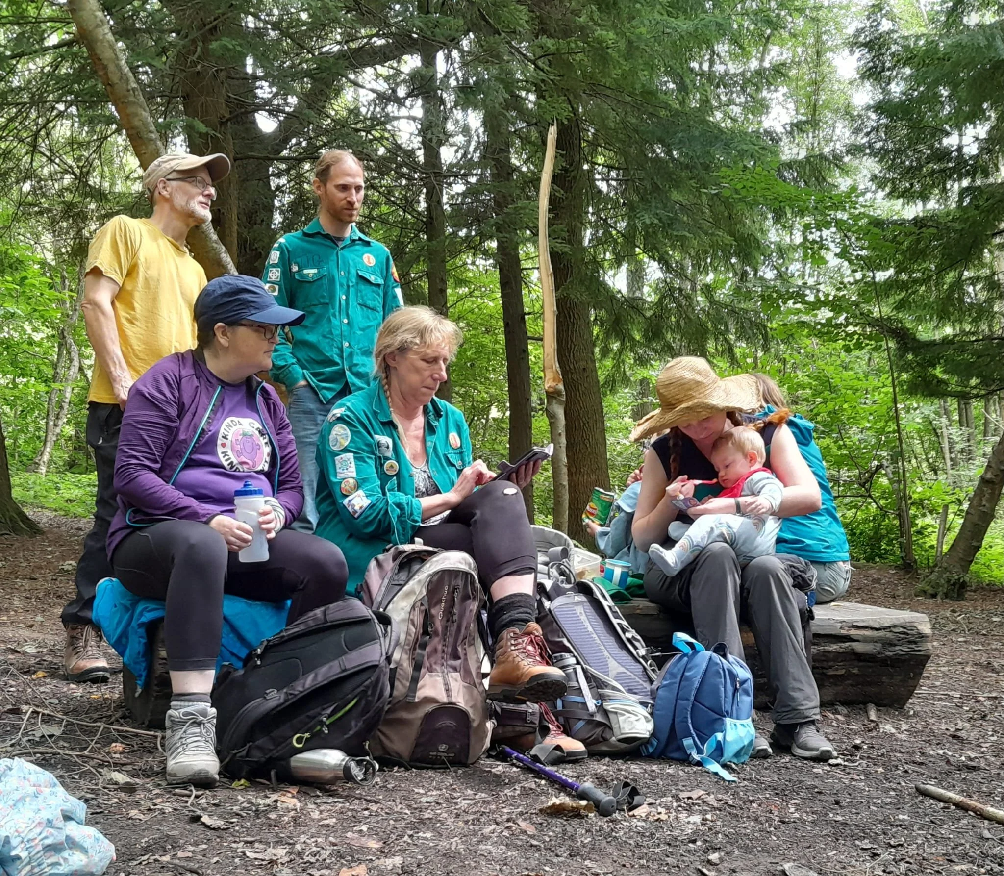 Group of people sitting and standing in a wooded outdoor area, surrounded by trees and forest, with backpacks and outdoor gear.