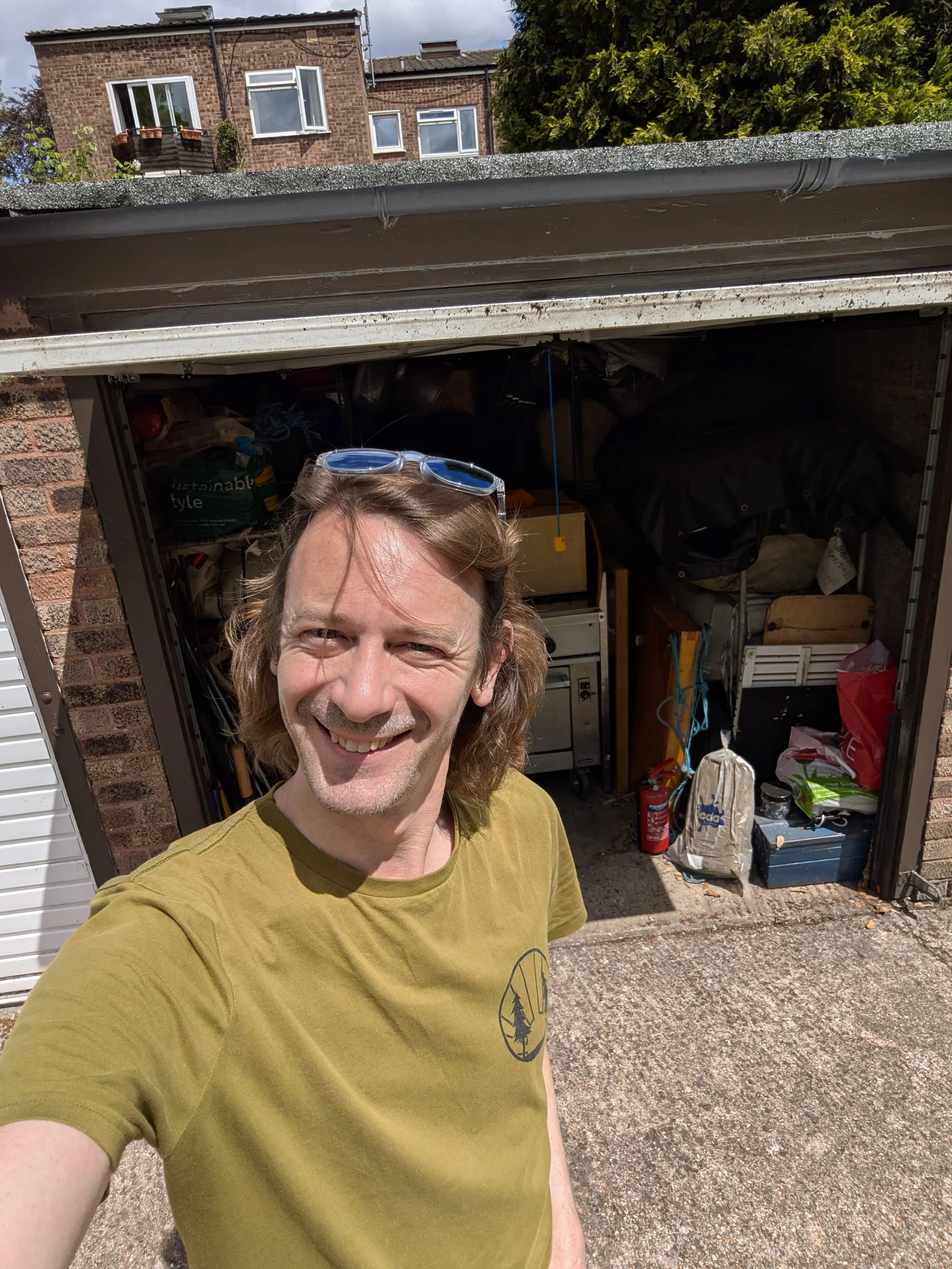 A smiling man with long hair and sunglasses on his head taking a selfie outside a garage filled with stored items.