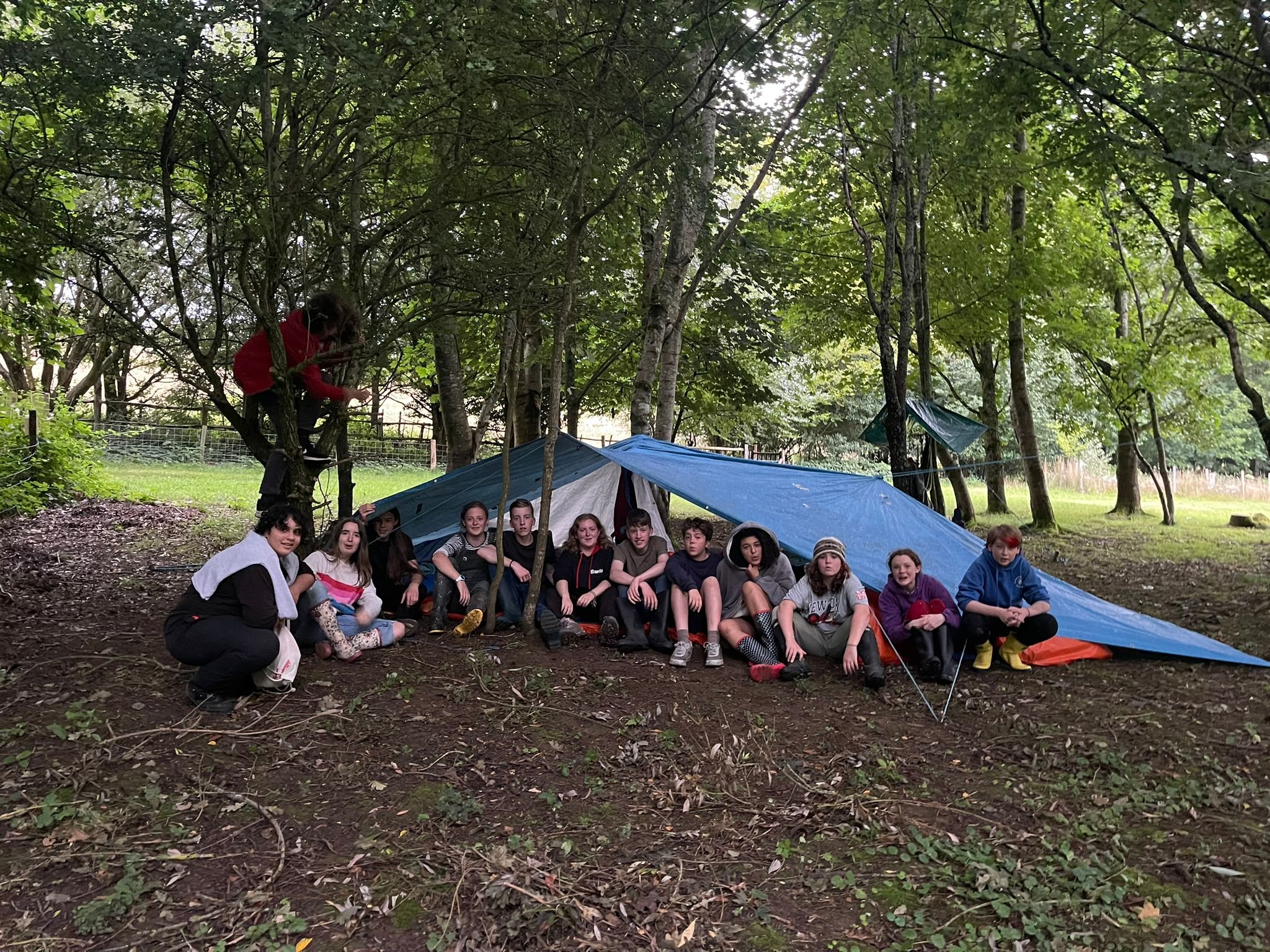 A group of children and a counselor sitting under a blue tarp in a wooded area, possibly on a camping trip, with trees and a fence in the background.