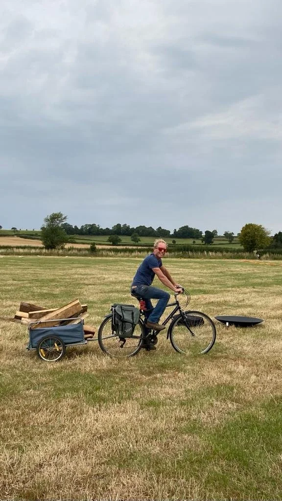 Man riding a bicycle in a grassy field, pulling a small wagon loaded with firewood, with a cloudy sky overhead.