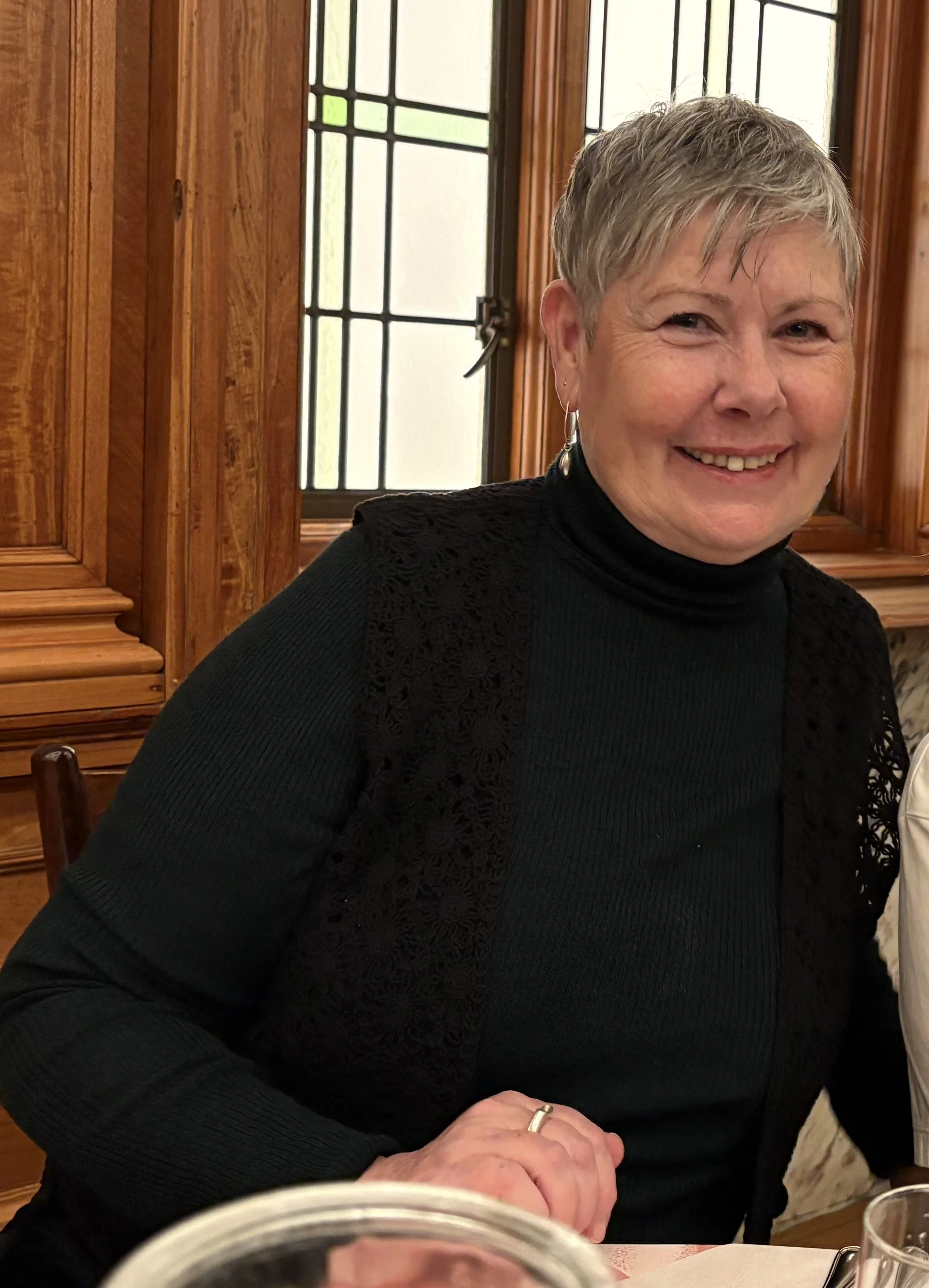 A smiling woman with short gray hair wearing a black top with lace details, sitting at a table inside a warmly lit room with wooden paneling and stained glass windows.