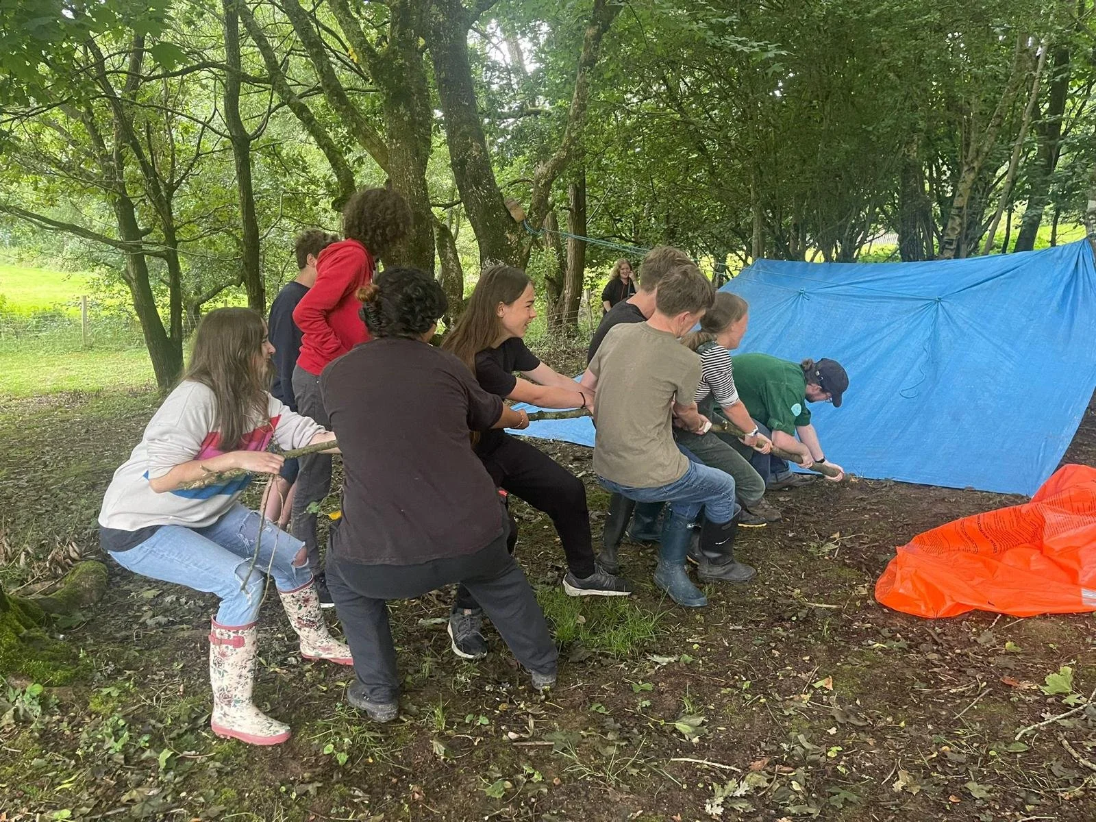 A group of people participating in a team-building activity outdoors in a wooded area. They are pulling on a rope, working together and smiling near a blue tarp and orange material on the ground.