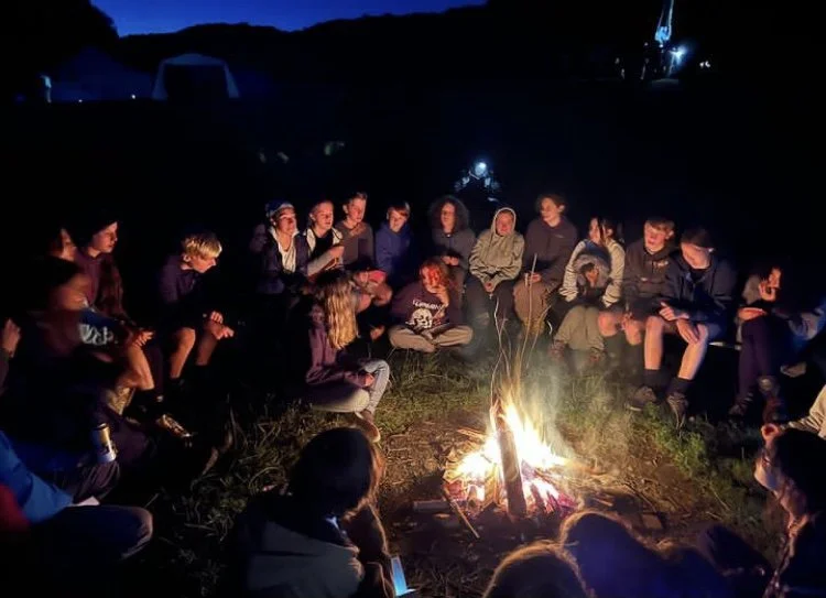 Group of people gathered around a campfire outdoors at night, sitting on the ground and enjoying each other's company.