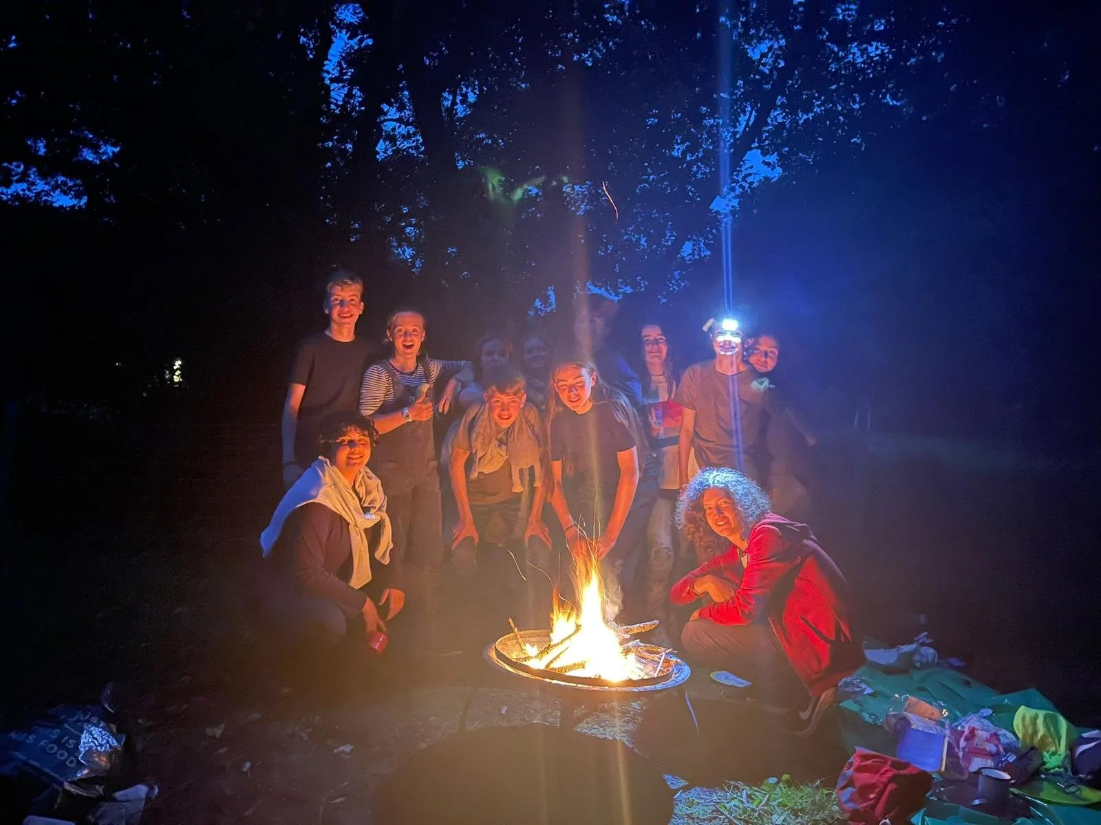 A group of people gathered around a campfire at night, sitting and standing outdoors, with smiles and cheerful expressions, surrounded by trees and some scattered camping gear.