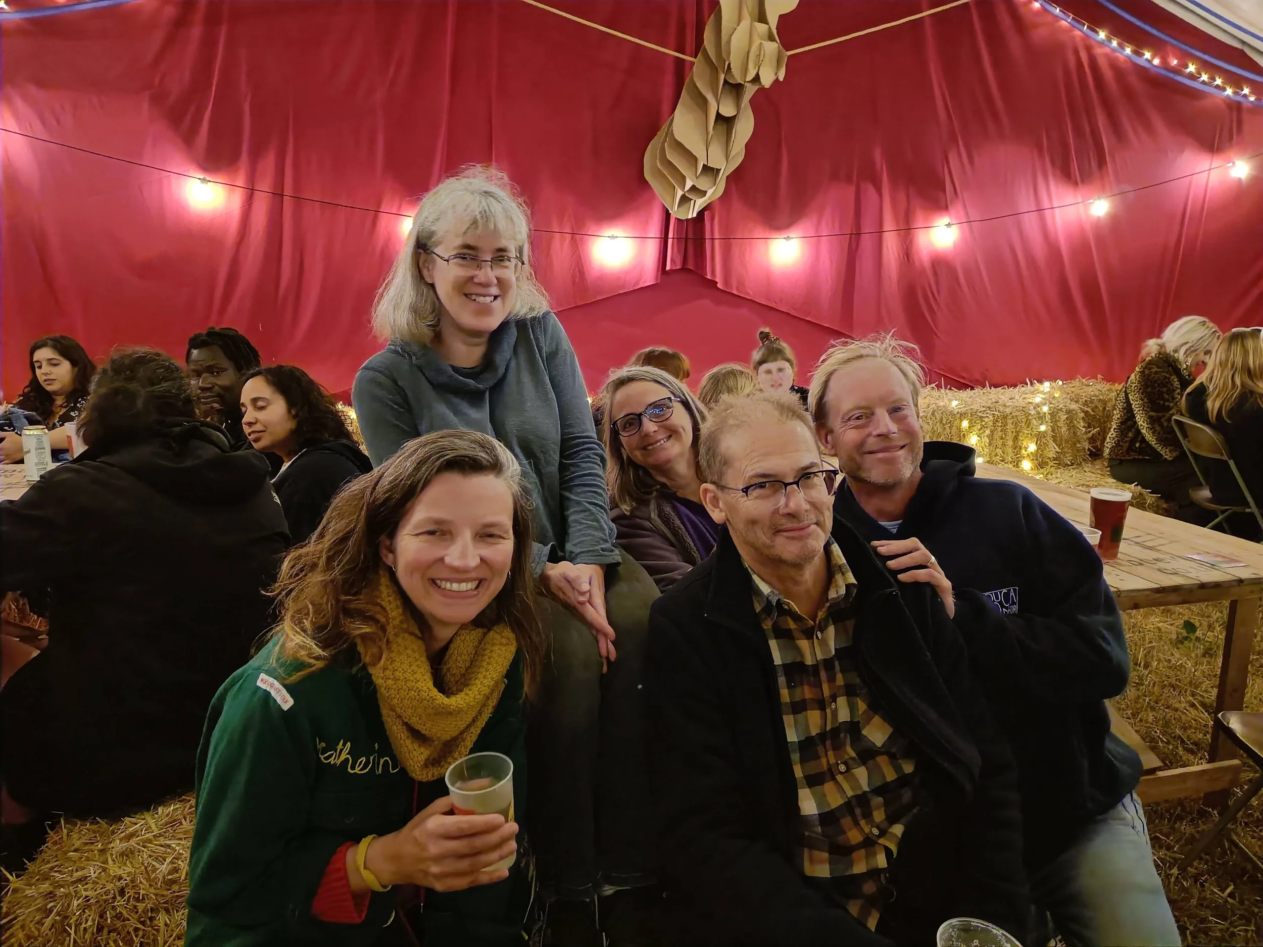 People sitting and smiling at a social gathering inside a decorated tent with string lights, red curtains, and hay on the ground.