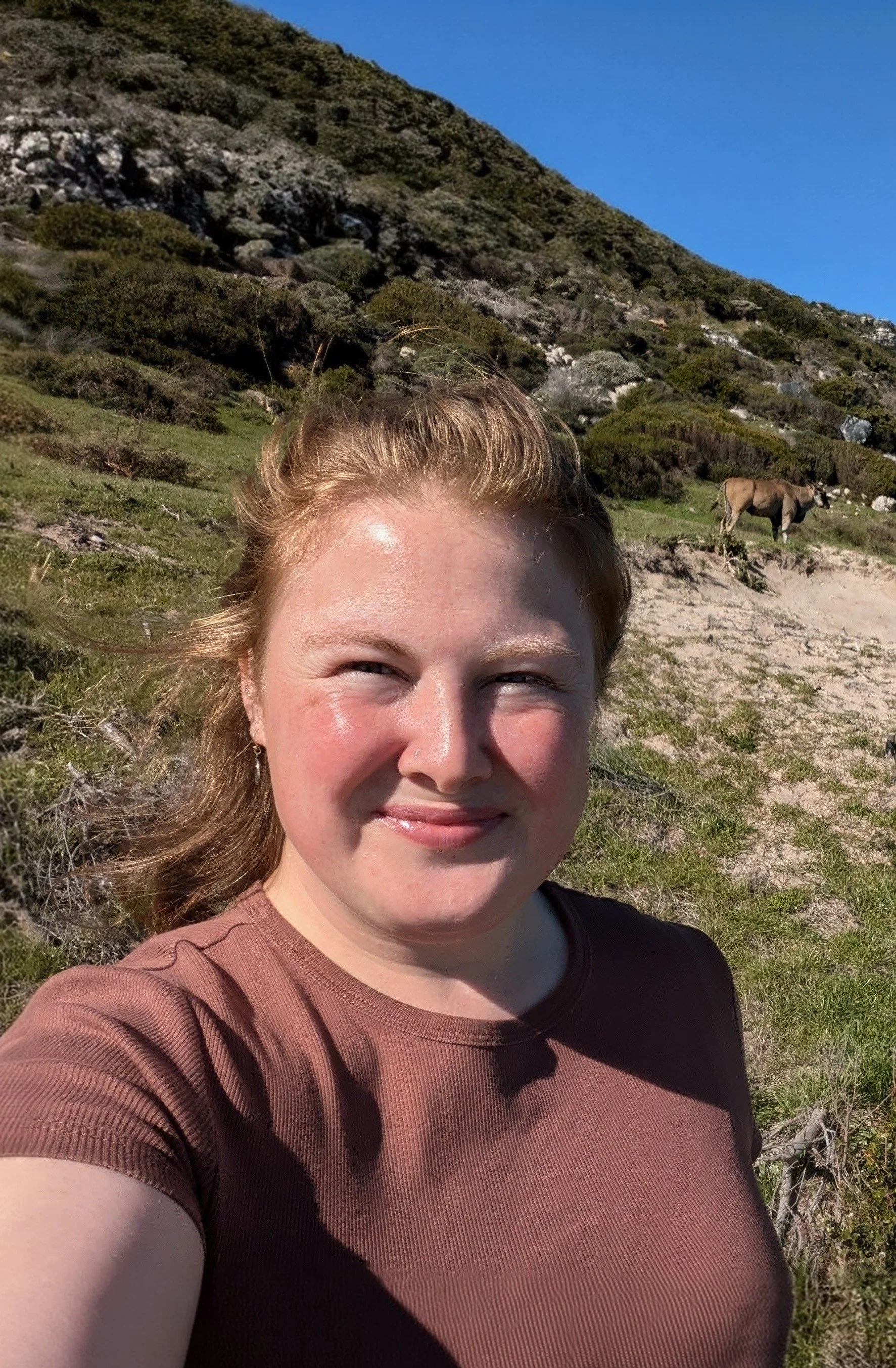 Close-up of a smiling woman with red hair taking a selfie outdoors on a sunny day. In the background, there is a mountain with green vegetation, a horse, and a clear blue sky.