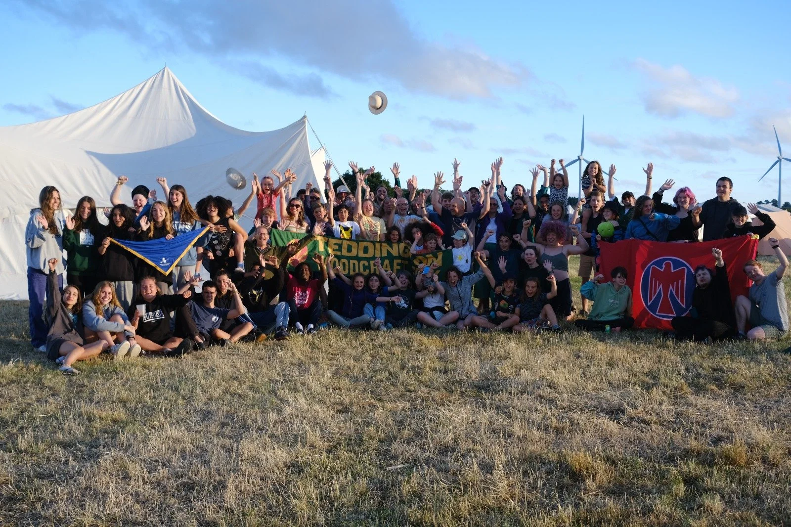 A large group of people gathered outdoors on a grassy field, raising their hands and smiling. Some are holding flags and banners with text and logos, and there is a large white tent and wind turbines in the background.