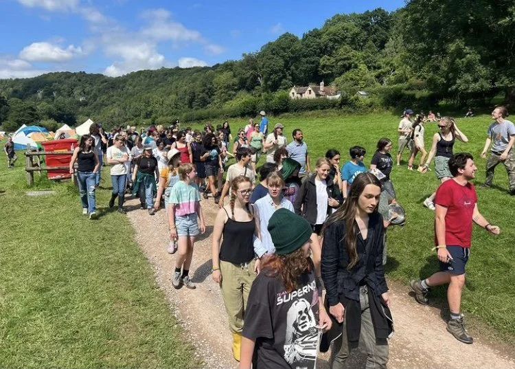 Large group of people walking along a dirt path in a park with green grass and trees, some tents in the background, and houses on a hillside under a blue sky.