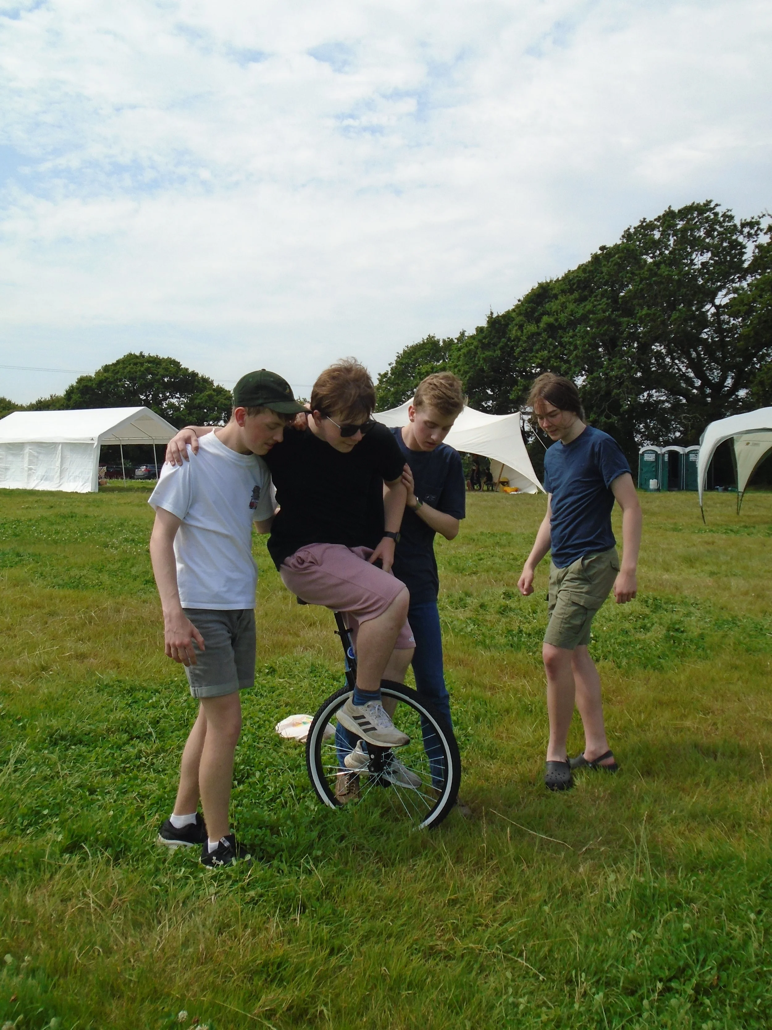 Four young people standing on grass in a park, one of them balancing on a unicycle while the others support him, with tents and trees in the background.