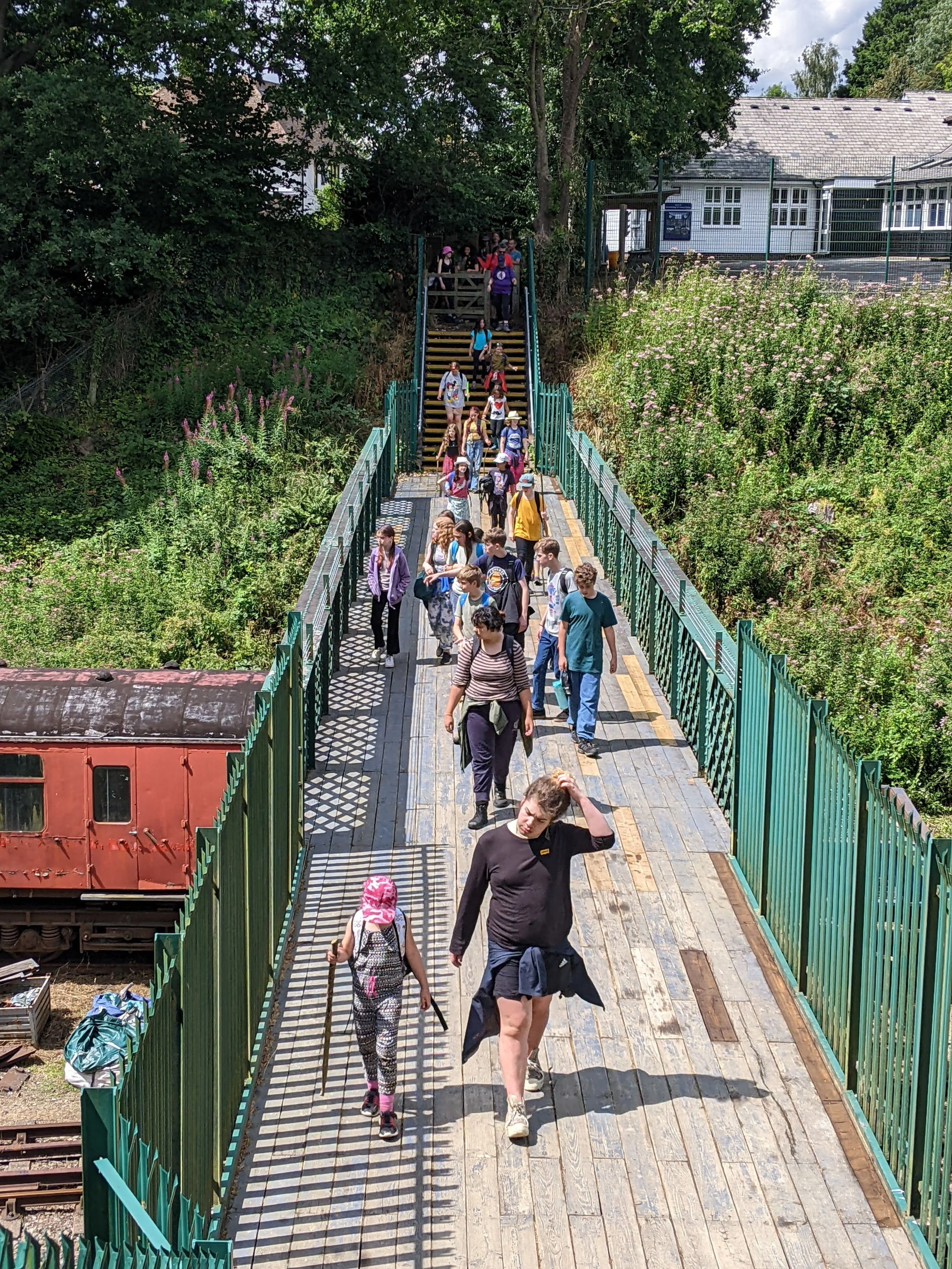 A group of people, including children, walking down a wooden bridge with metal railings, surrounded by greenery and trees, with a building and railway tracks visible in the background.