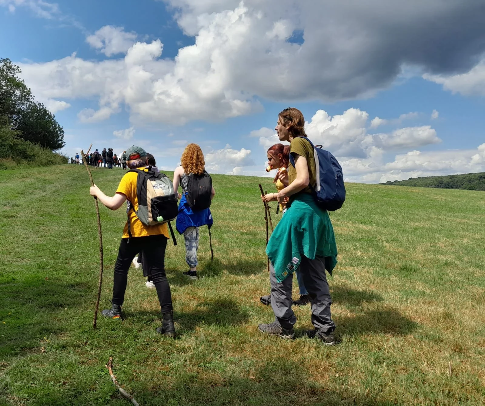 Hikers walking up a grassy hill on a partly cloudy day, with a group visible in the background.