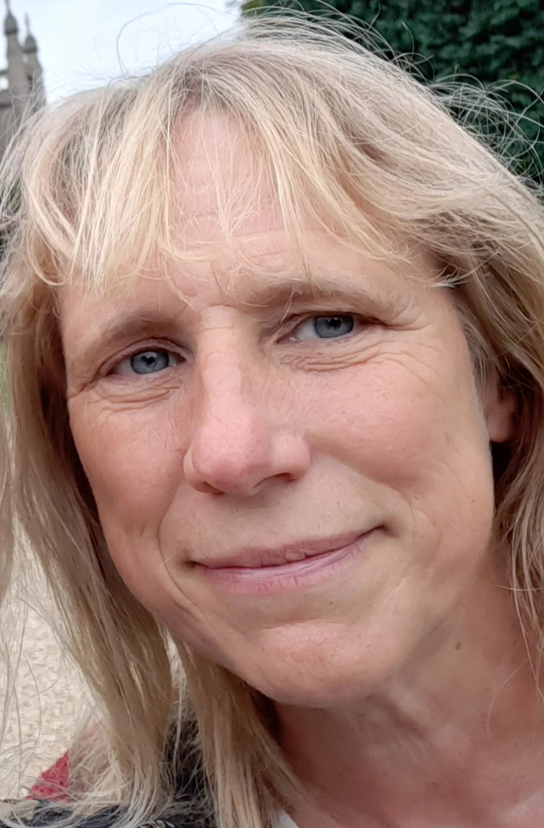 Close-up of a smiling middle-aged woman with blonde hair, blue eyes, and visible fine lines, outdoors with a blurred green background.