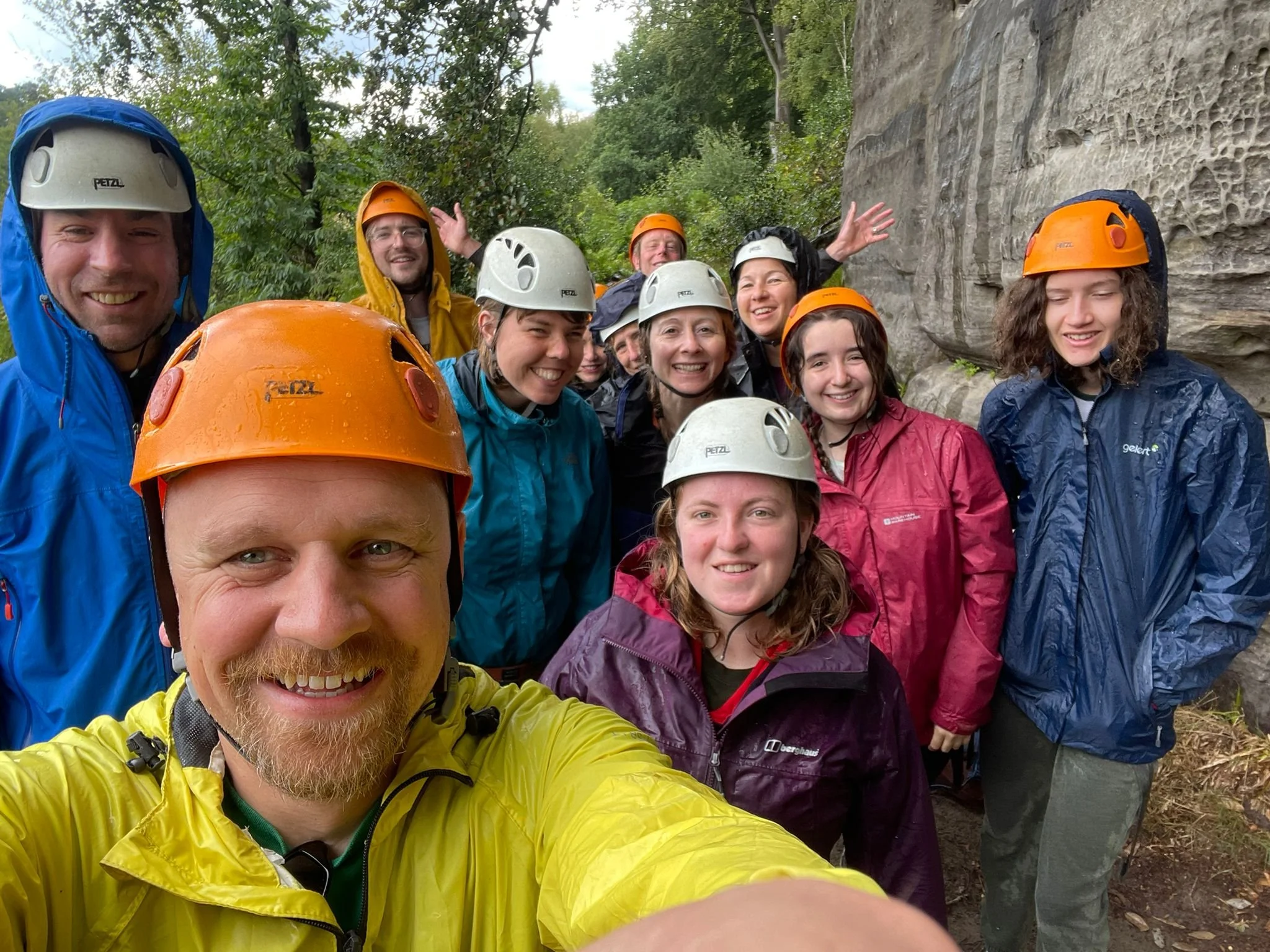 Group of people rock climbing outdoors, wearing helmets and rain jackets, smiling for a selfie with a rock face in the background.